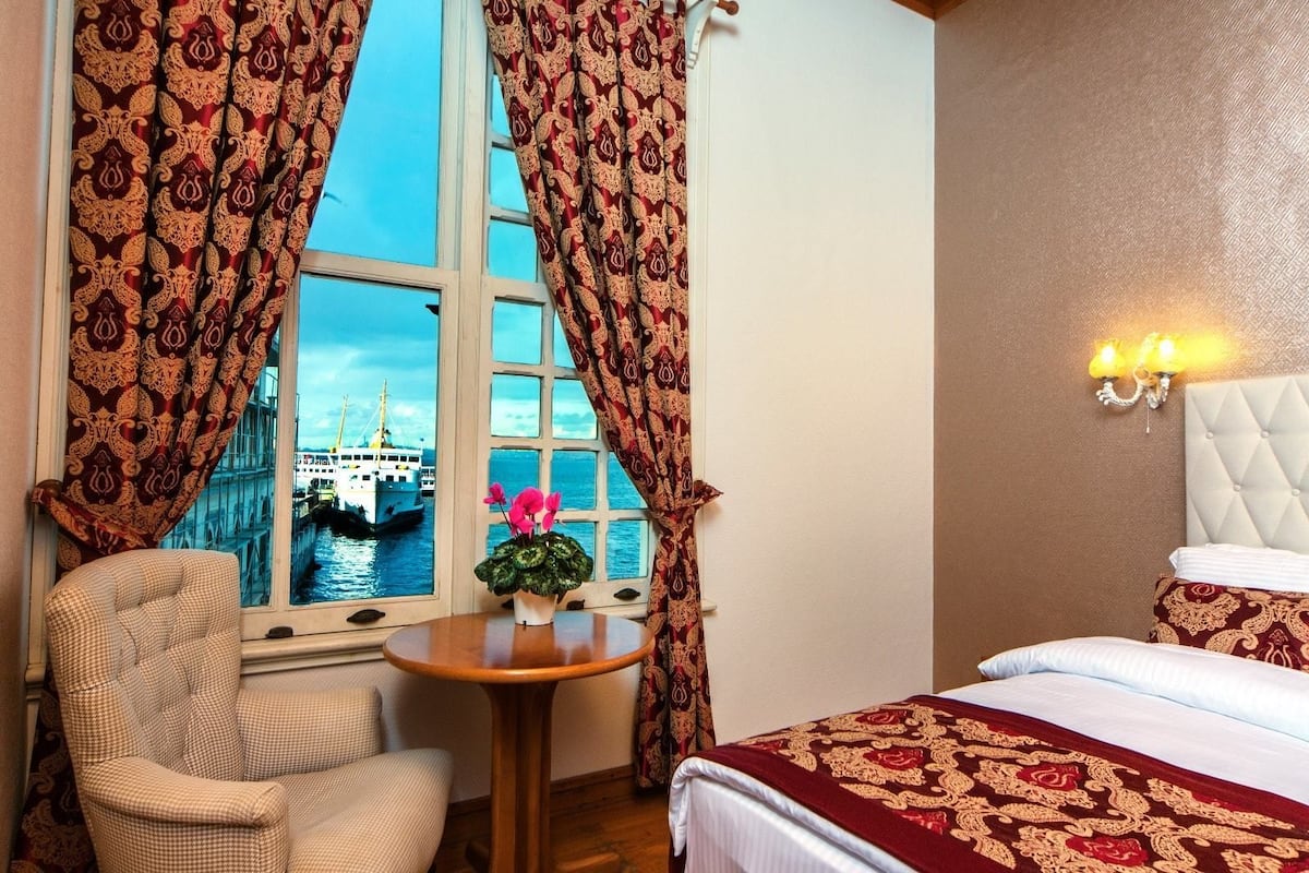 A hotel room with a large window, a view of a boat, a bed, a chair, and a small table with a plant.