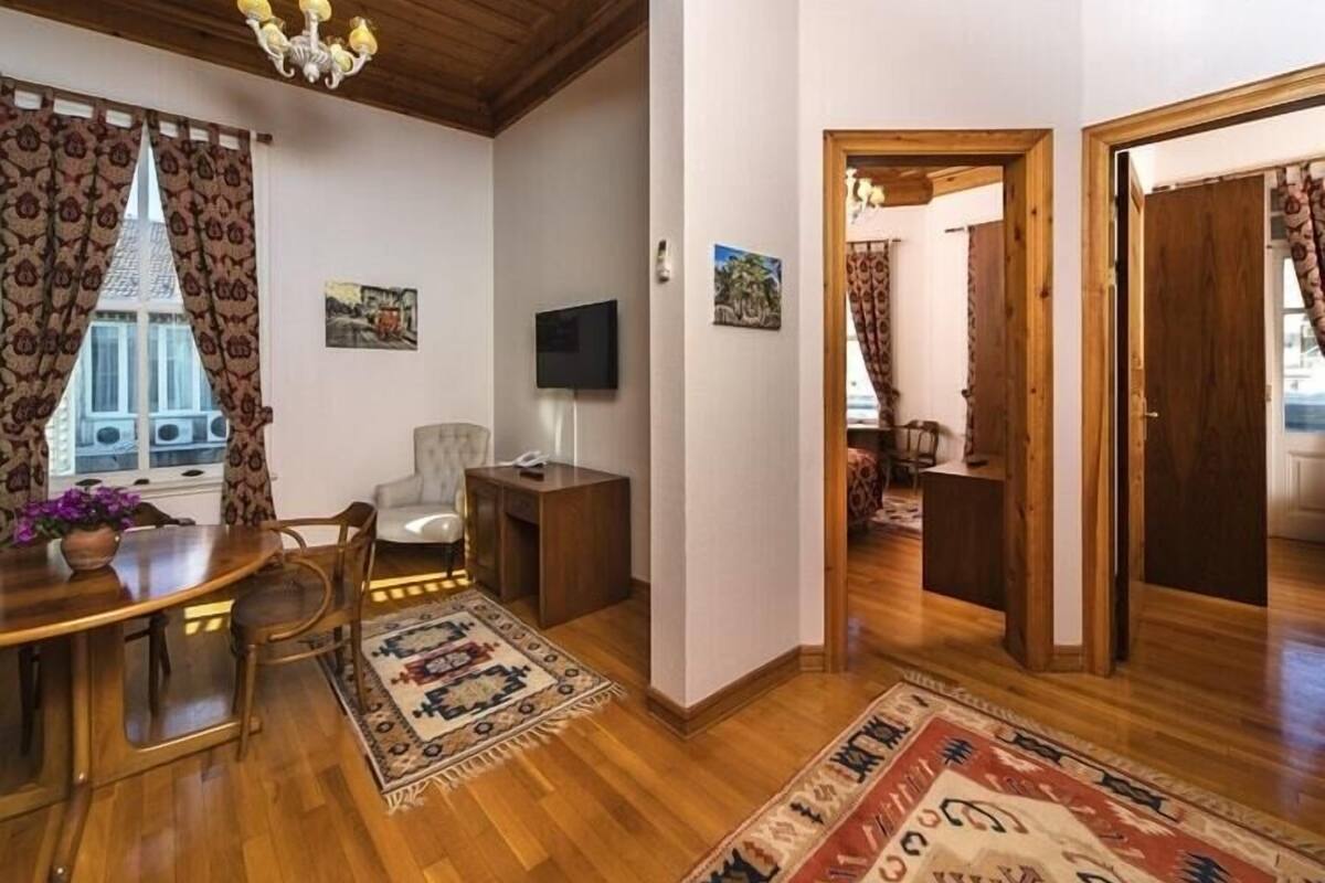 A room with a wooden ceiling, a chandelier, a dining table with chairs, a TV, and a patterned rug.