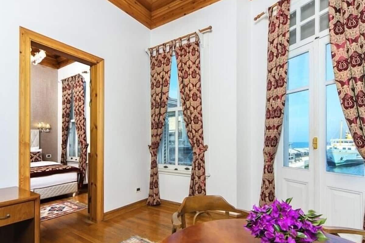 A room with a bed, a wooden desk, a dining table with a vase of flowers, patterned curtains, and a view of the sea through large windows.