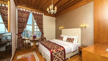 A hotel room with a wooden ceiling, a bed with patterned bedding, a desk with a chair, a small table with a vase, and a view of the city through large windows.