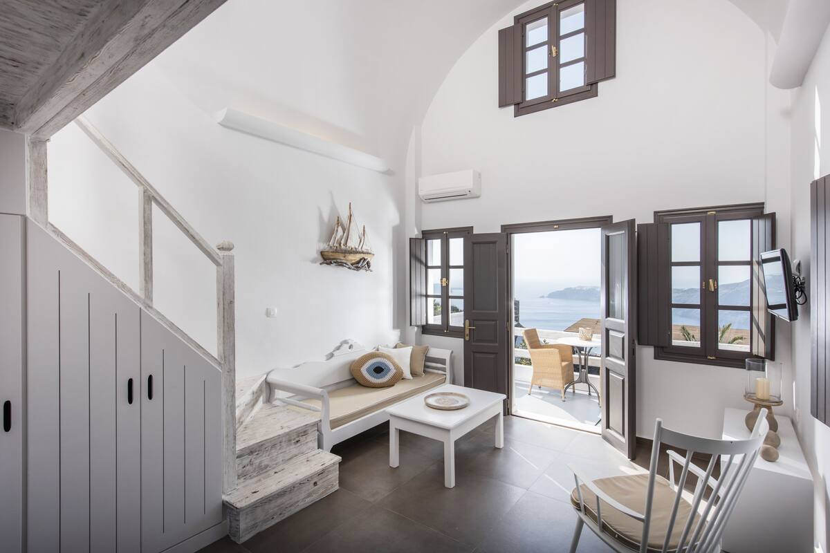 A spacious room with a white sofa, a small table, and a chair. There is a staircase leading to another level, and a door that opens to a balcony with a view of the sea.
