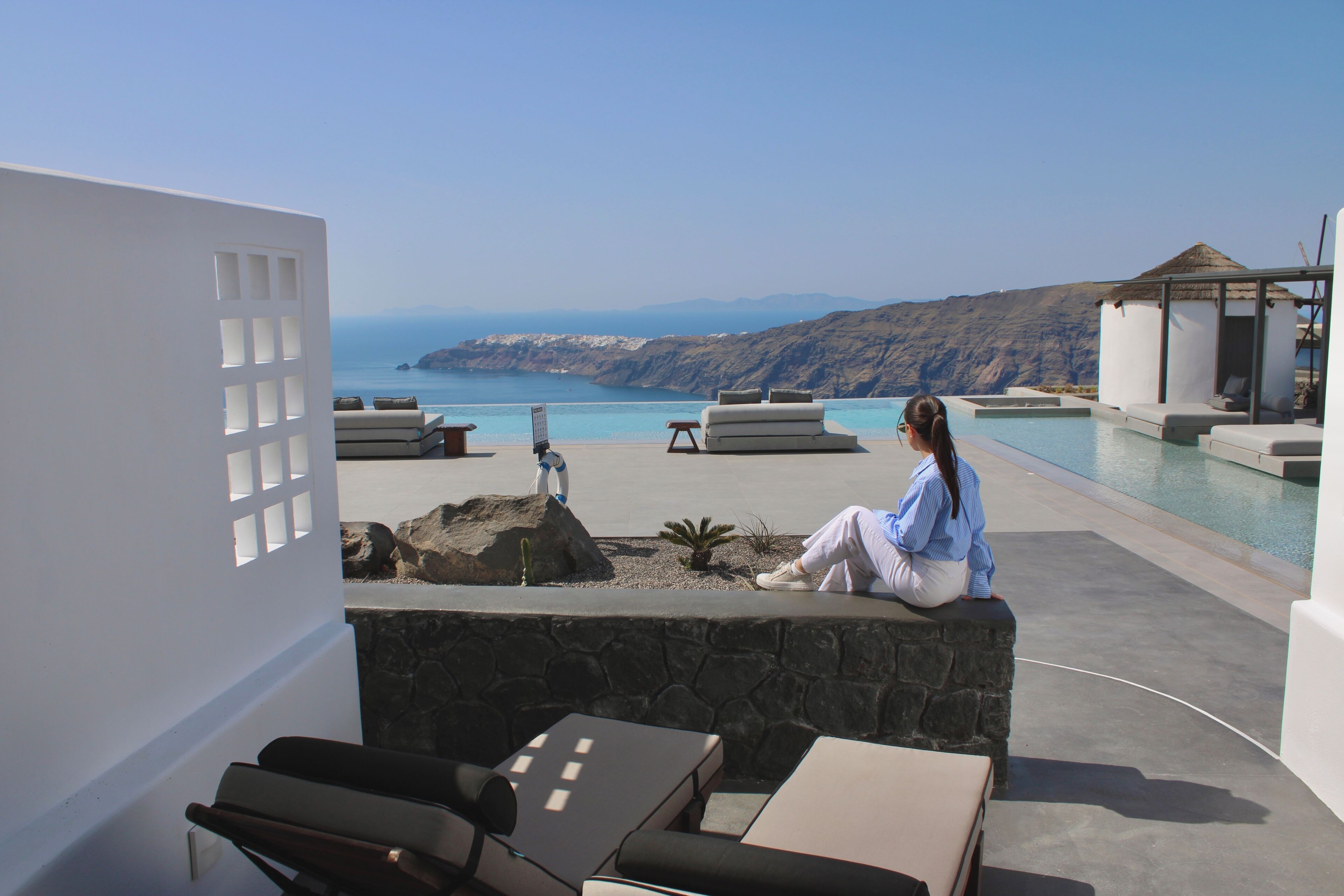 A poolside area with lounge chairs, a stone wall, and a view of the sea and mountains.