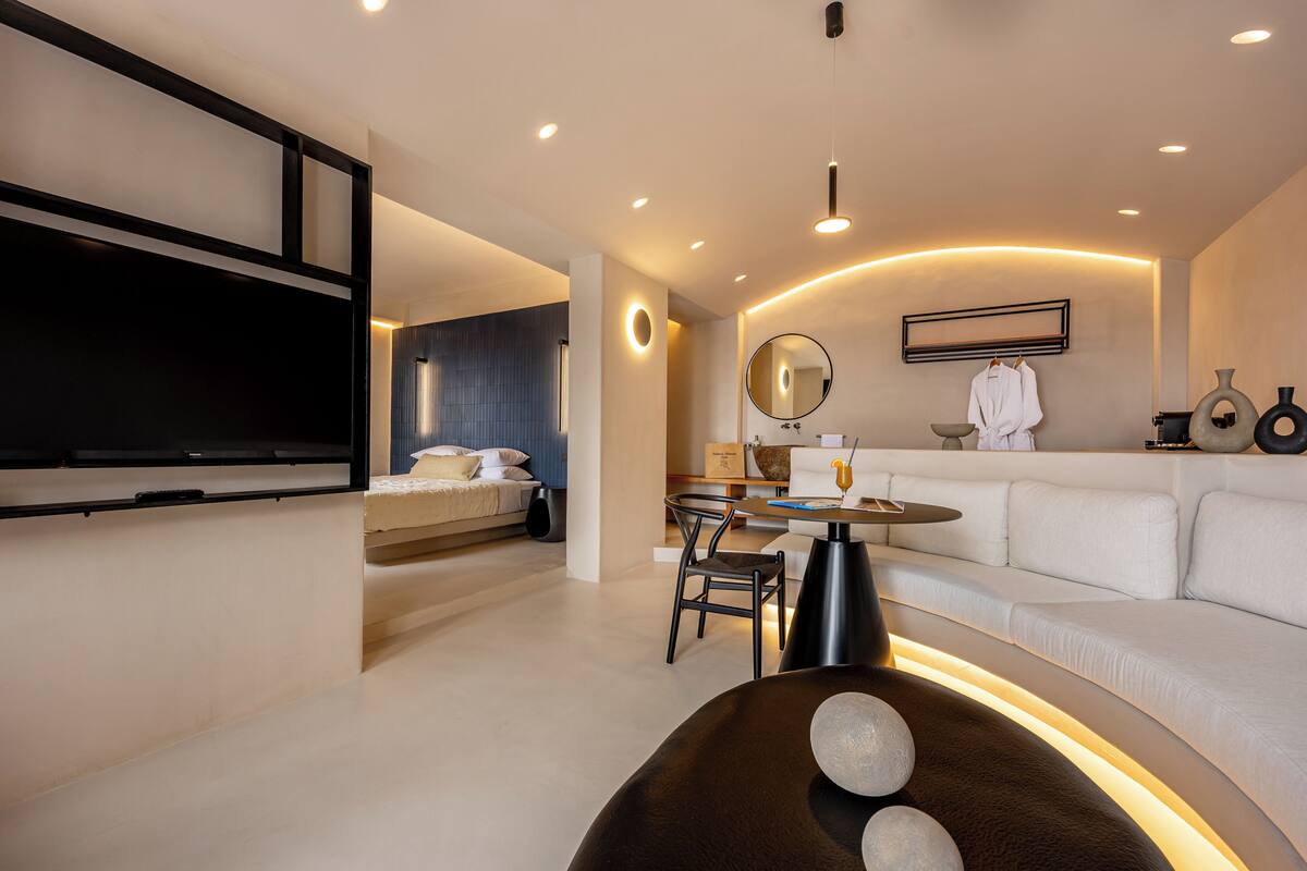 A modern hotel room with a large bed, a dining area, a sofa, and a TV mounted on the wall.