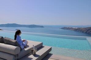 Una persona descansando en un sofá junto a una piscina infinita, con vistas despejadas al mar y a formaciones terrestres distantes.