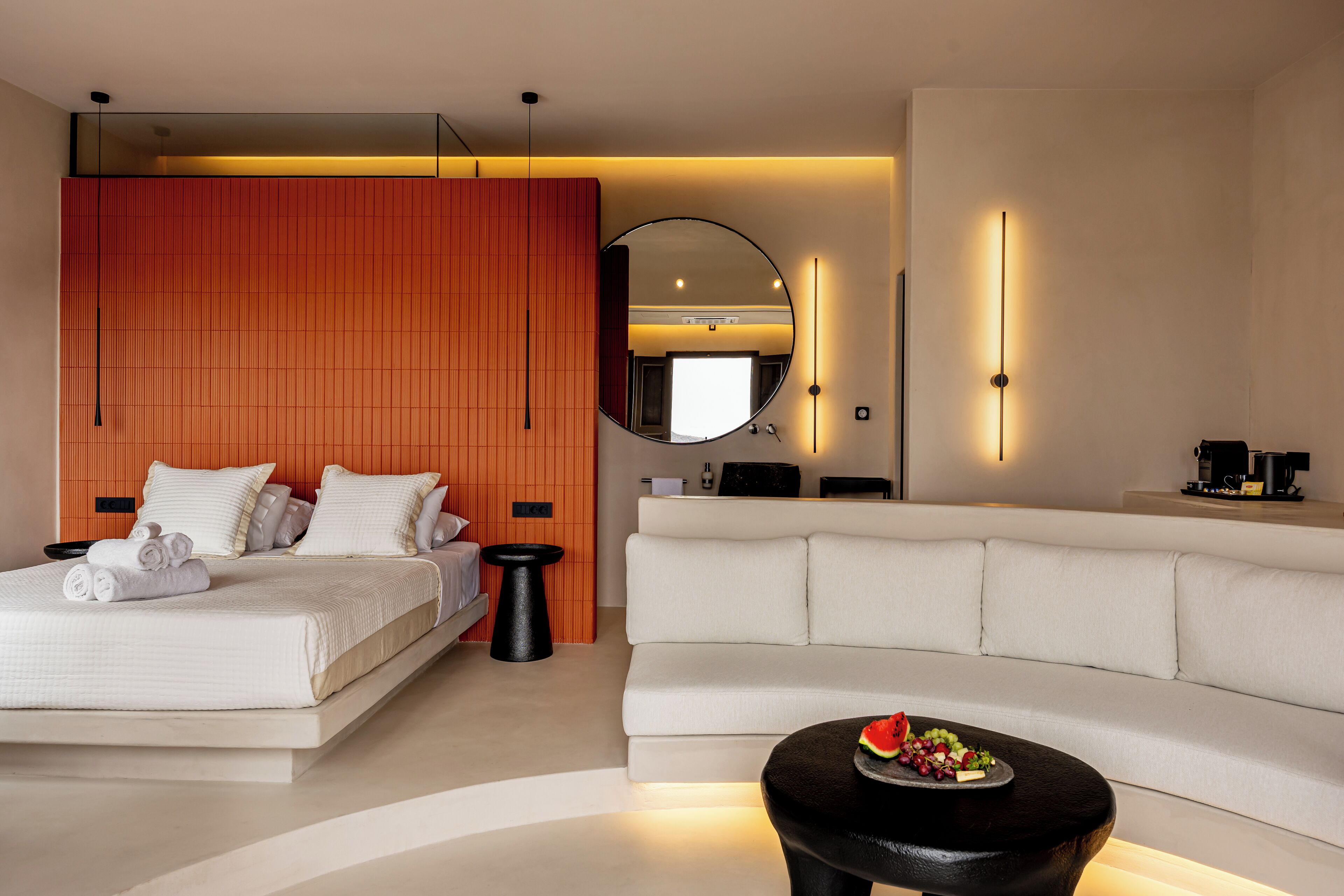 A modern hotel room with a bed, a white sofa, a round coffee table with fruit, and a large mirror.