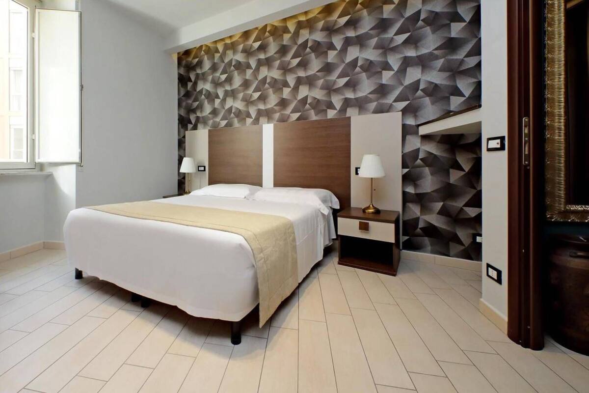 A modern hotel room with a large bed, a nightstand with a lamp, and a geometric-patterned wall.