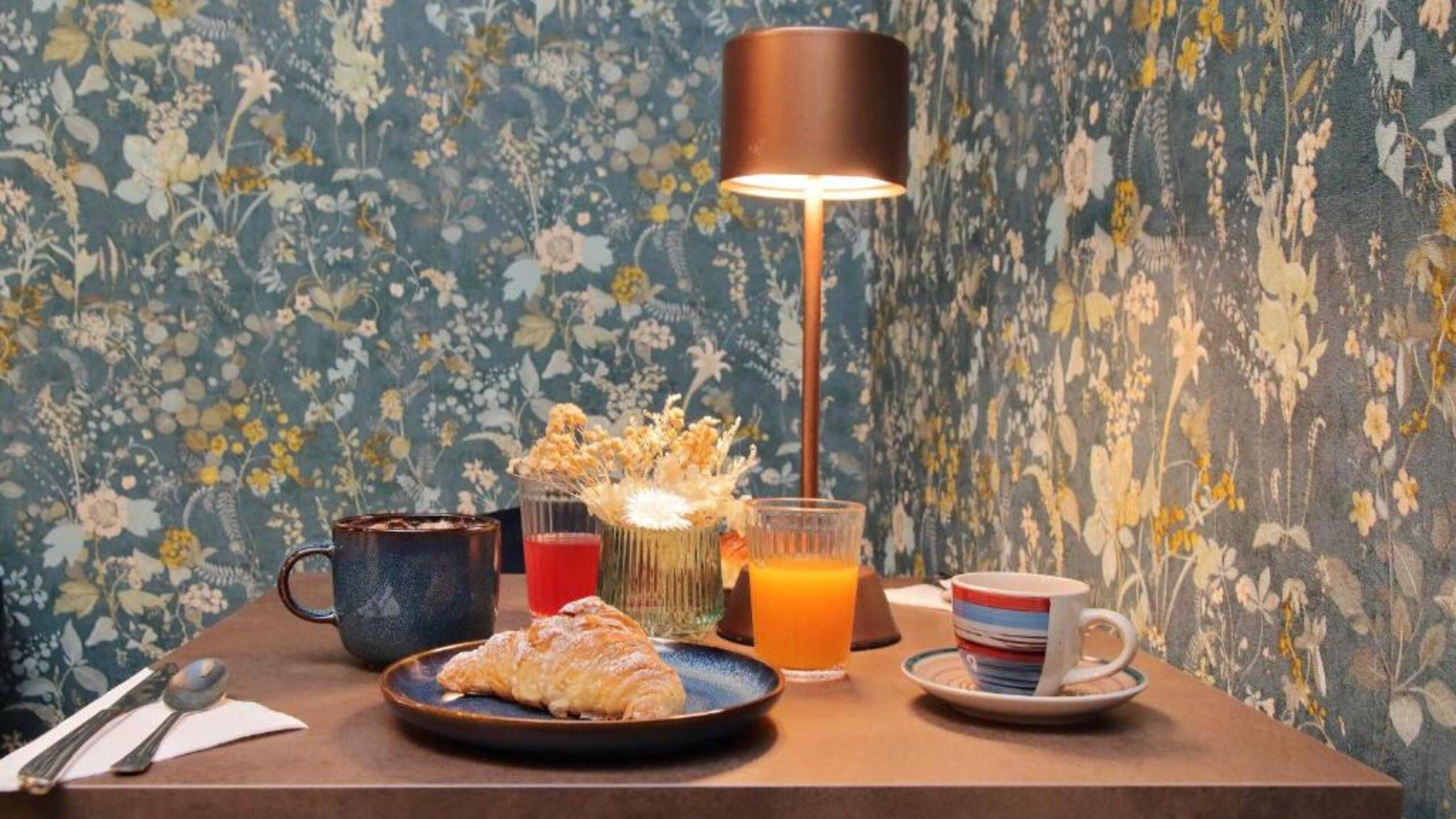 Una presentación para el desayuno con un croissant, una taza, un vaso de zumo de naranja y una taza sobre una mesa con un fondo de papel tapiz floral.