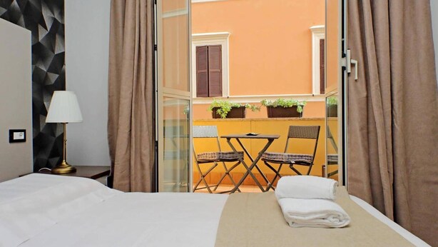 Een hotelkamer met een bed, een tafel met stoelen, een lamp en uitzicht op een balkon met planten en een raam.
