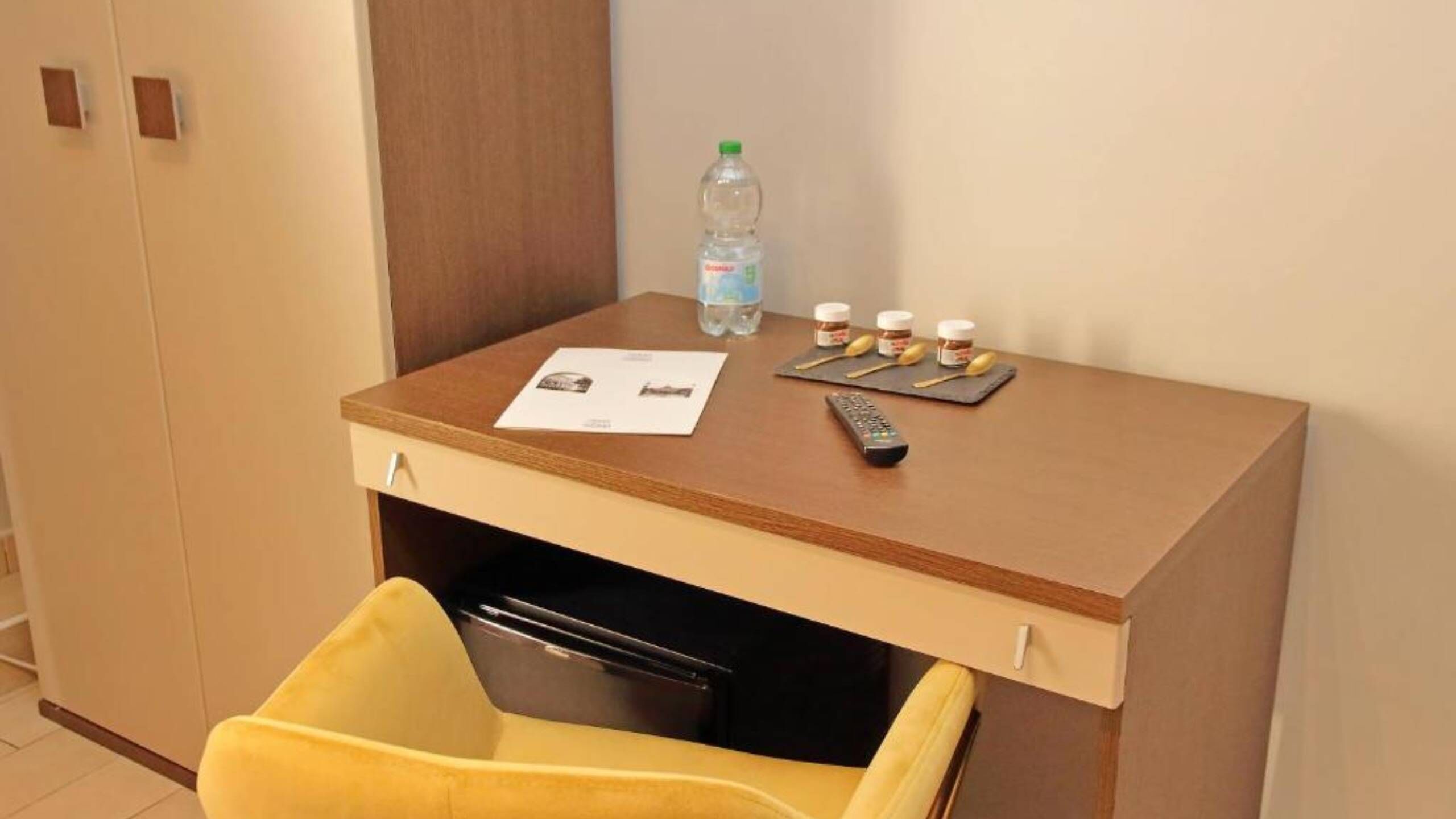 A compact hotel room with a wooden desk, a yellow chair, a remote control, a water bottle, and some snacks.