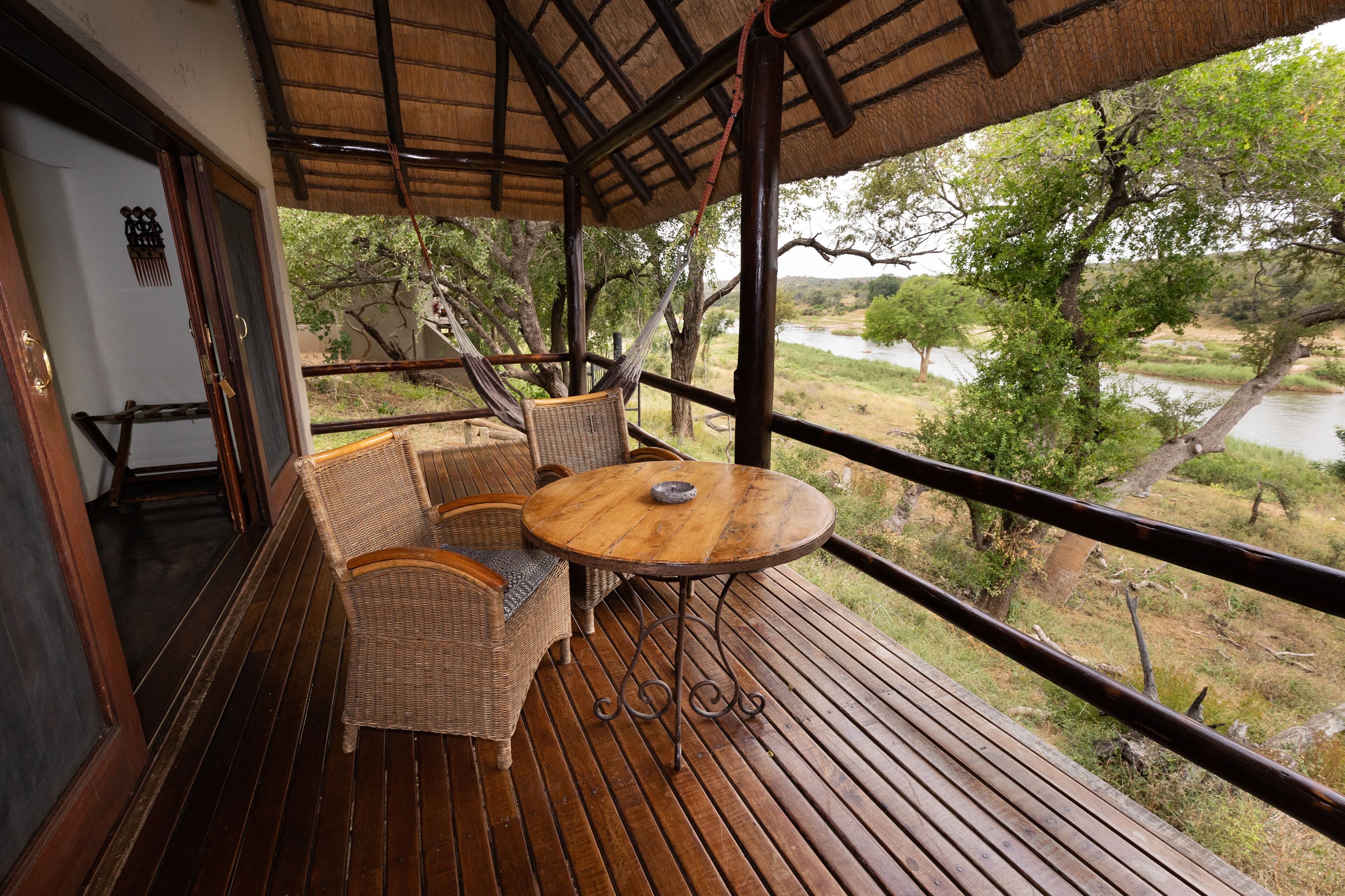 Terraza de madera con muebles de mimbre y una mesa, con vista a un río y árboles.