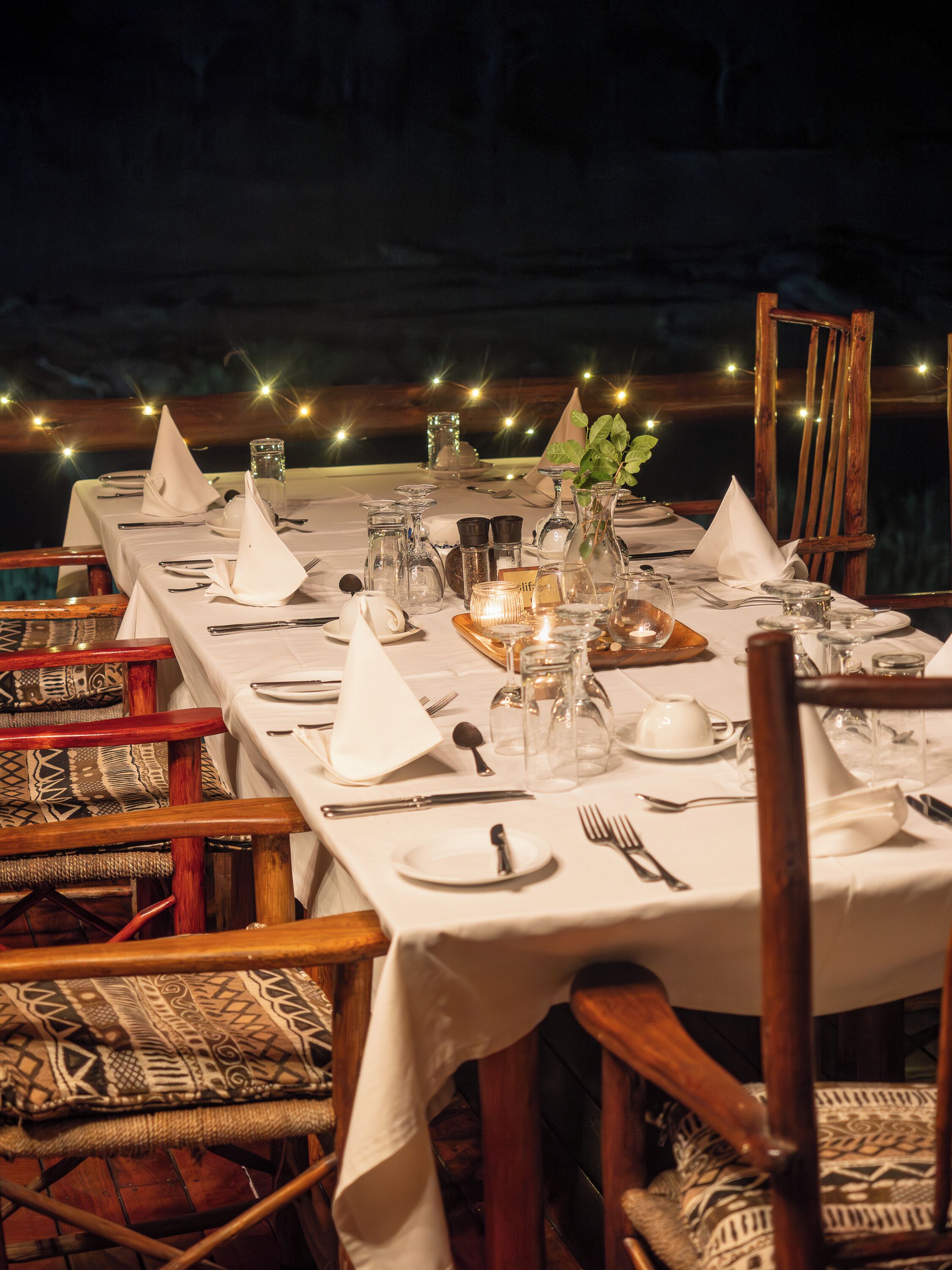 A table set for a meal with white tablecloths, glassware, and wooden chairs.