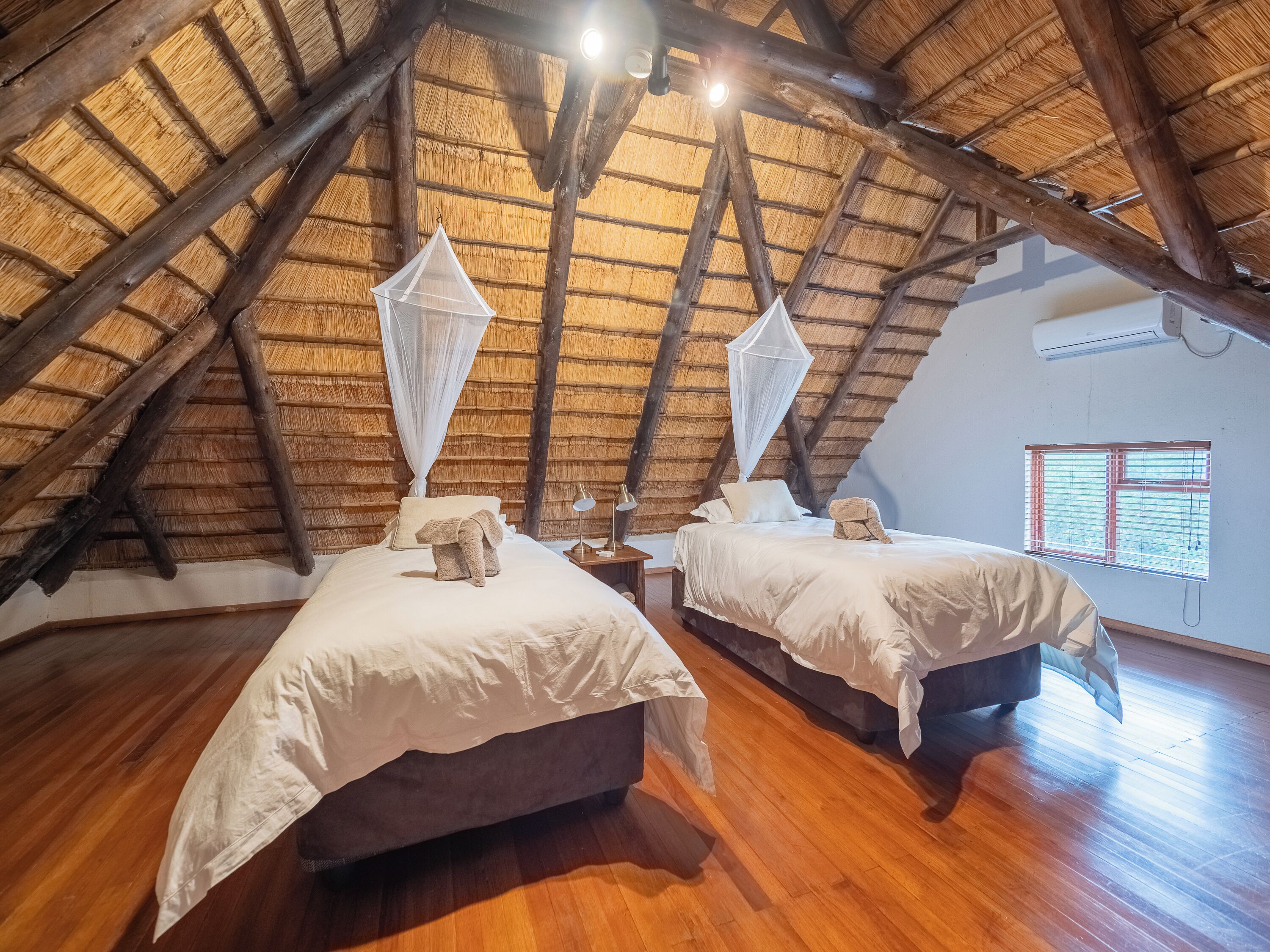 A room with two beds, a wooden floor, a thatched roof, and a window with blinds.