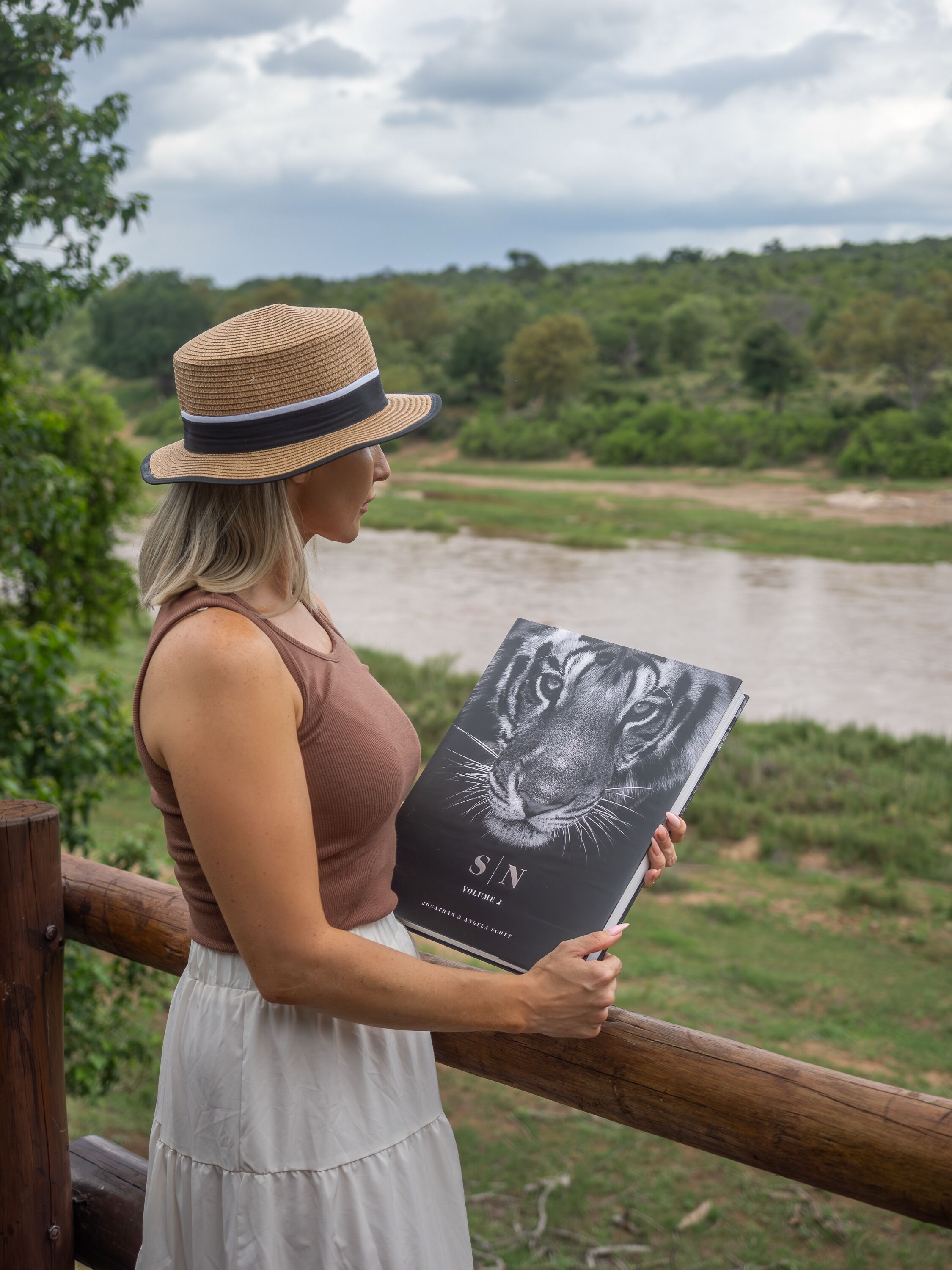A person holding a book titled "S/N Volume 2" by Joanna Inge and Angela Scott, standing by a wooden railing with a river and greenery in the background.
