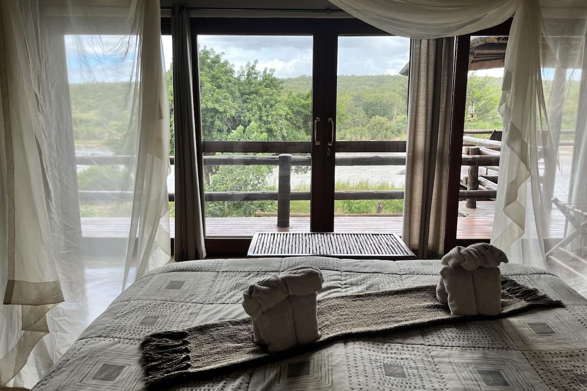 A room with a bed, a view of greenery, and a wooden deck outside.
