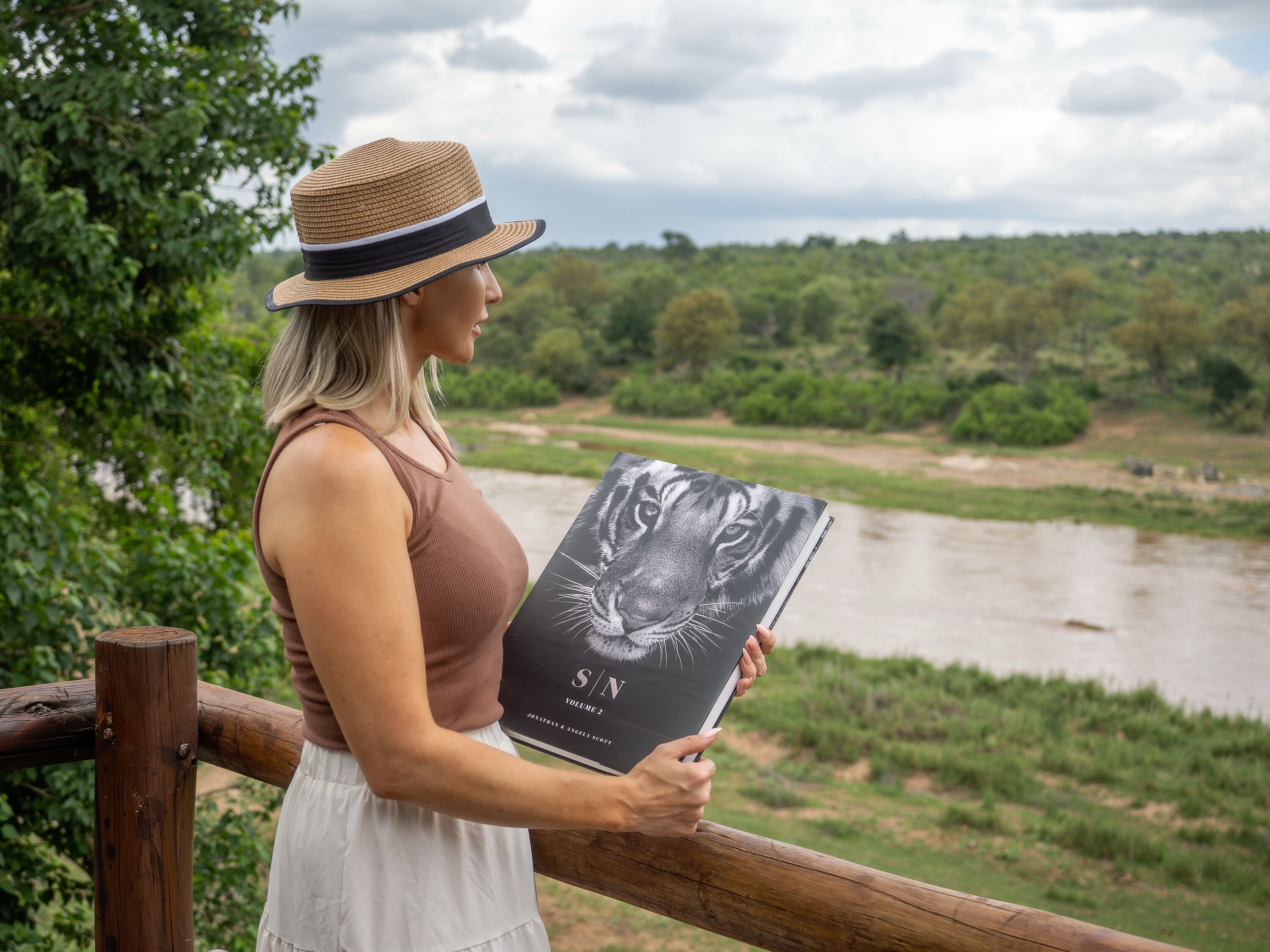 A person holding a book titled 'S/N Volume 2' by Joanna Scott and Angela Scott, standing by a wooden railing with a river and greenery in the background.