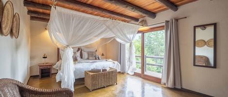 Un dormitorio con una cama con dosel, una silla de mimbre, una mesita de noche y un ventanal con vistas a zonas verdes.