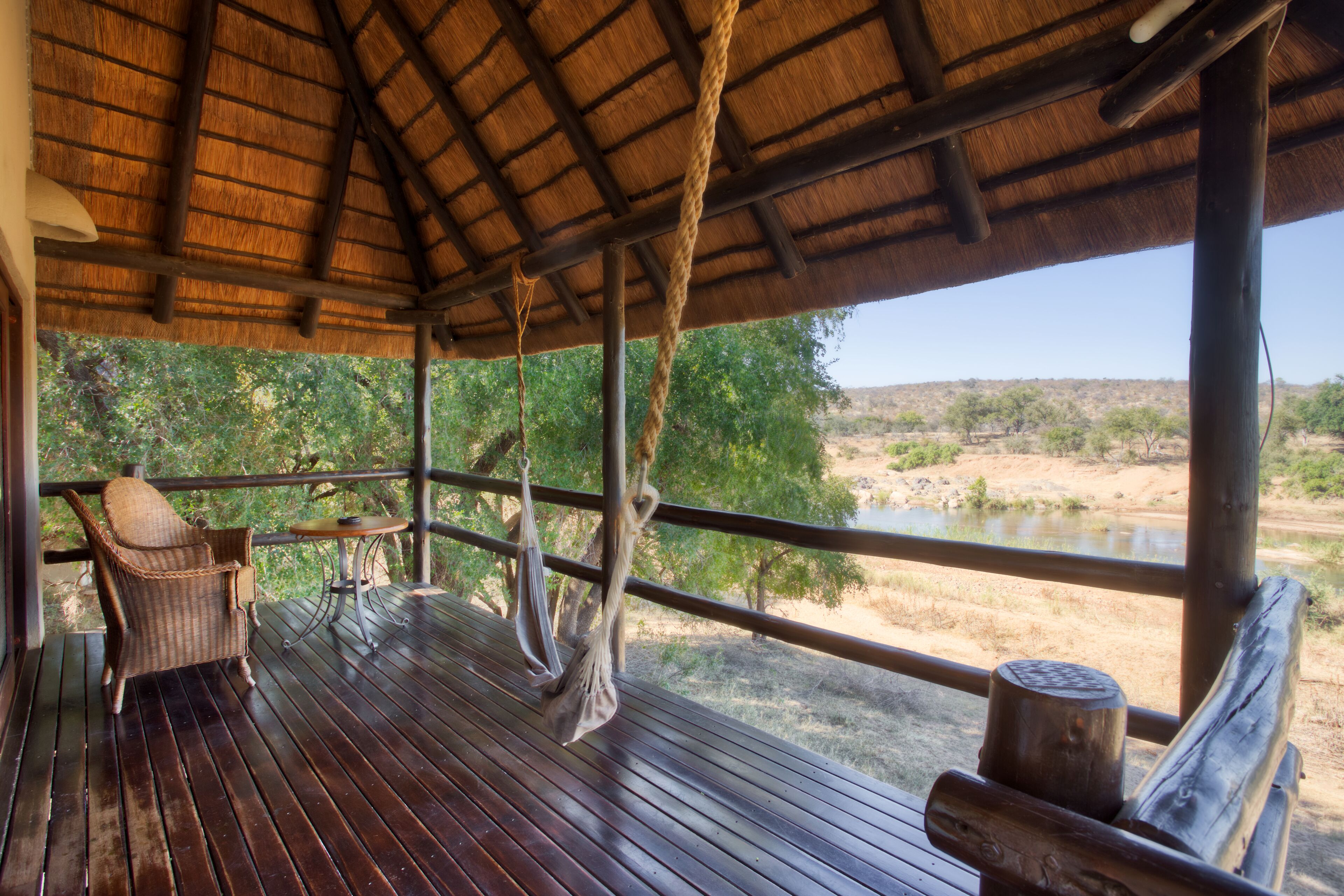 A wooden deck with a thatched roof, wicker furniture, and a view of a river and trees.
