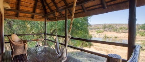 A wooden deck with a thatched roof, wicker furniture, and a view of a river and trees.