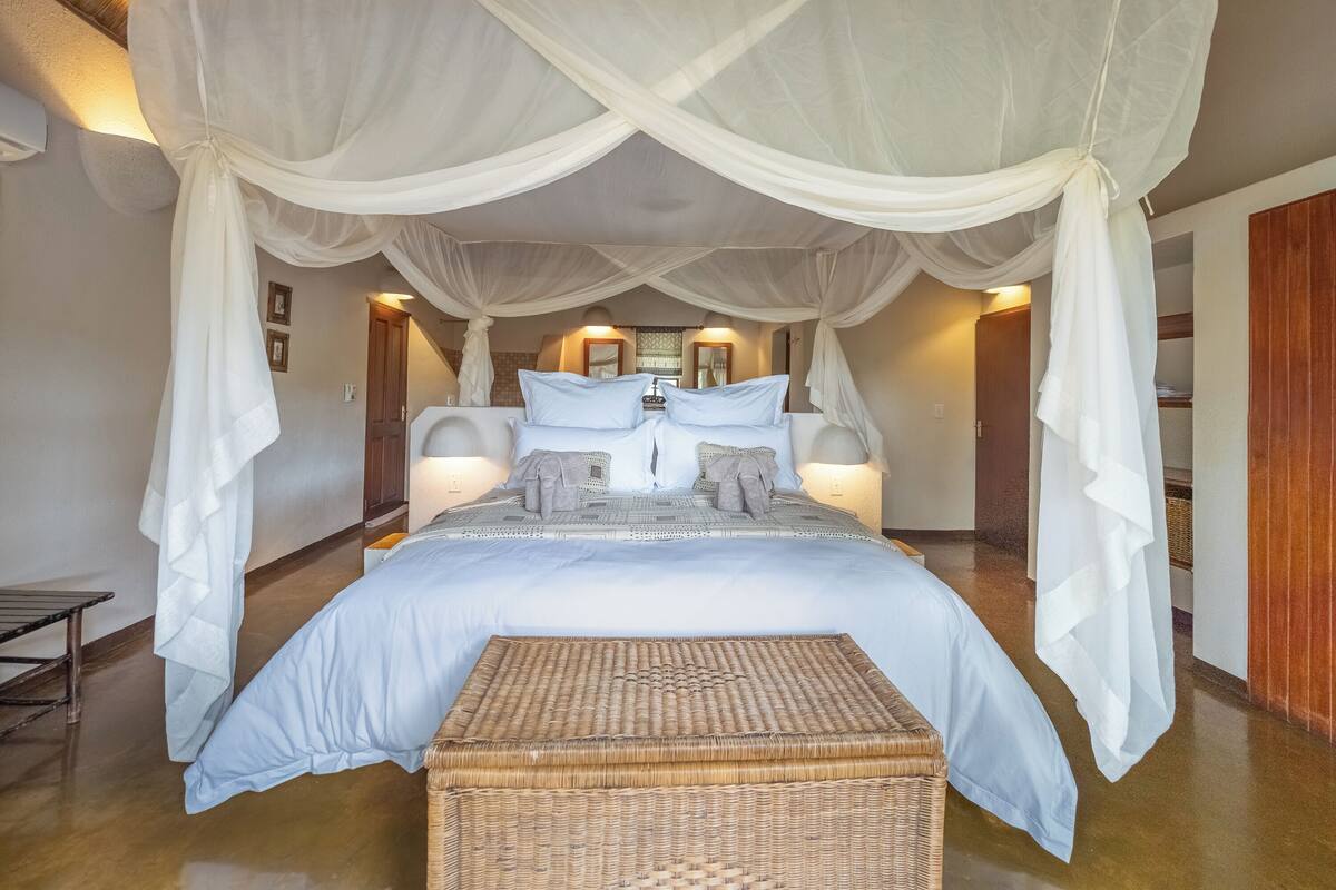 A bedroom with a canopy bed, bedside lamps, a wicker ottoman, and a wooden floor.