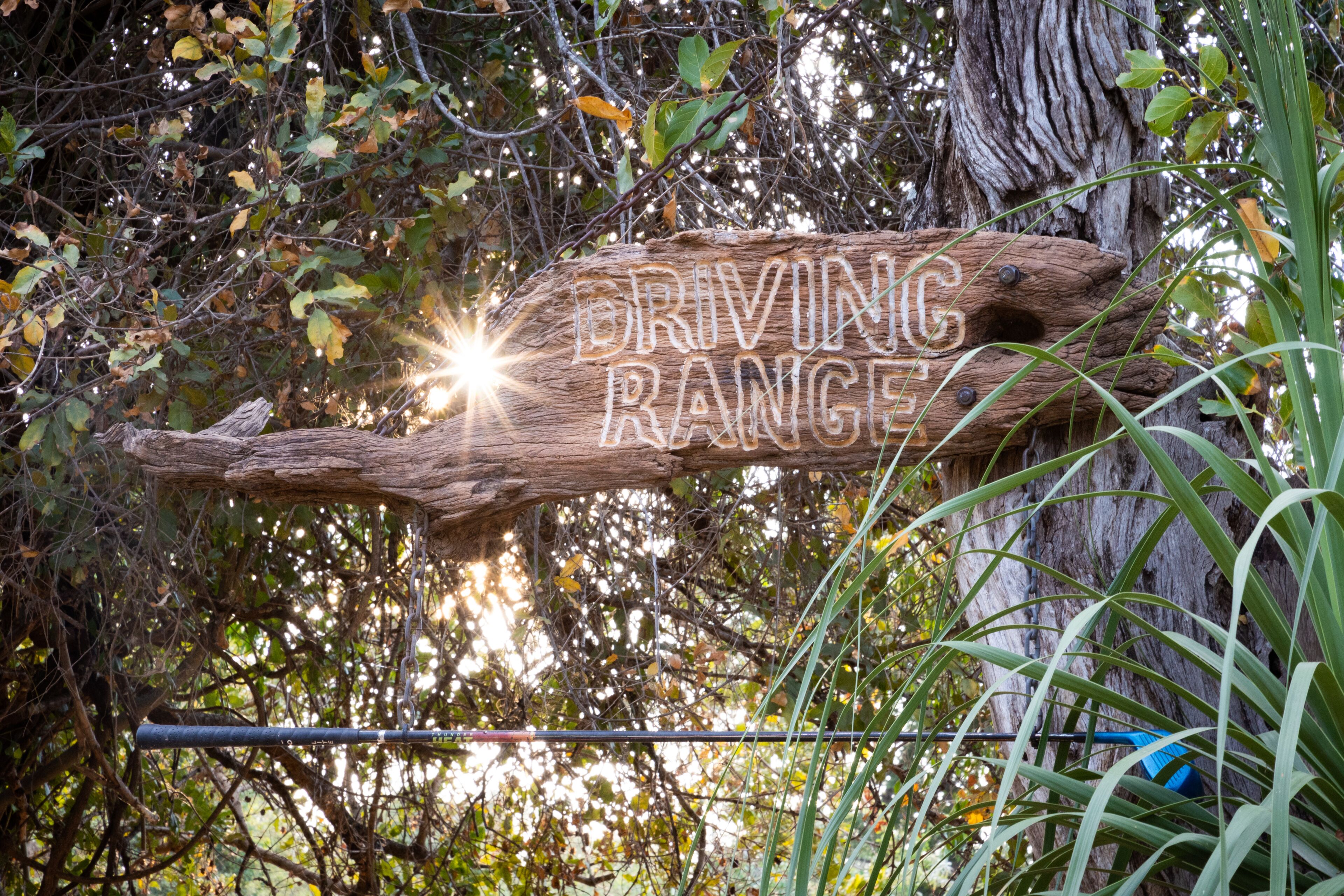 A wooden sign with the text "DRIVING RANGE" in a natural setting with trees and foliage.