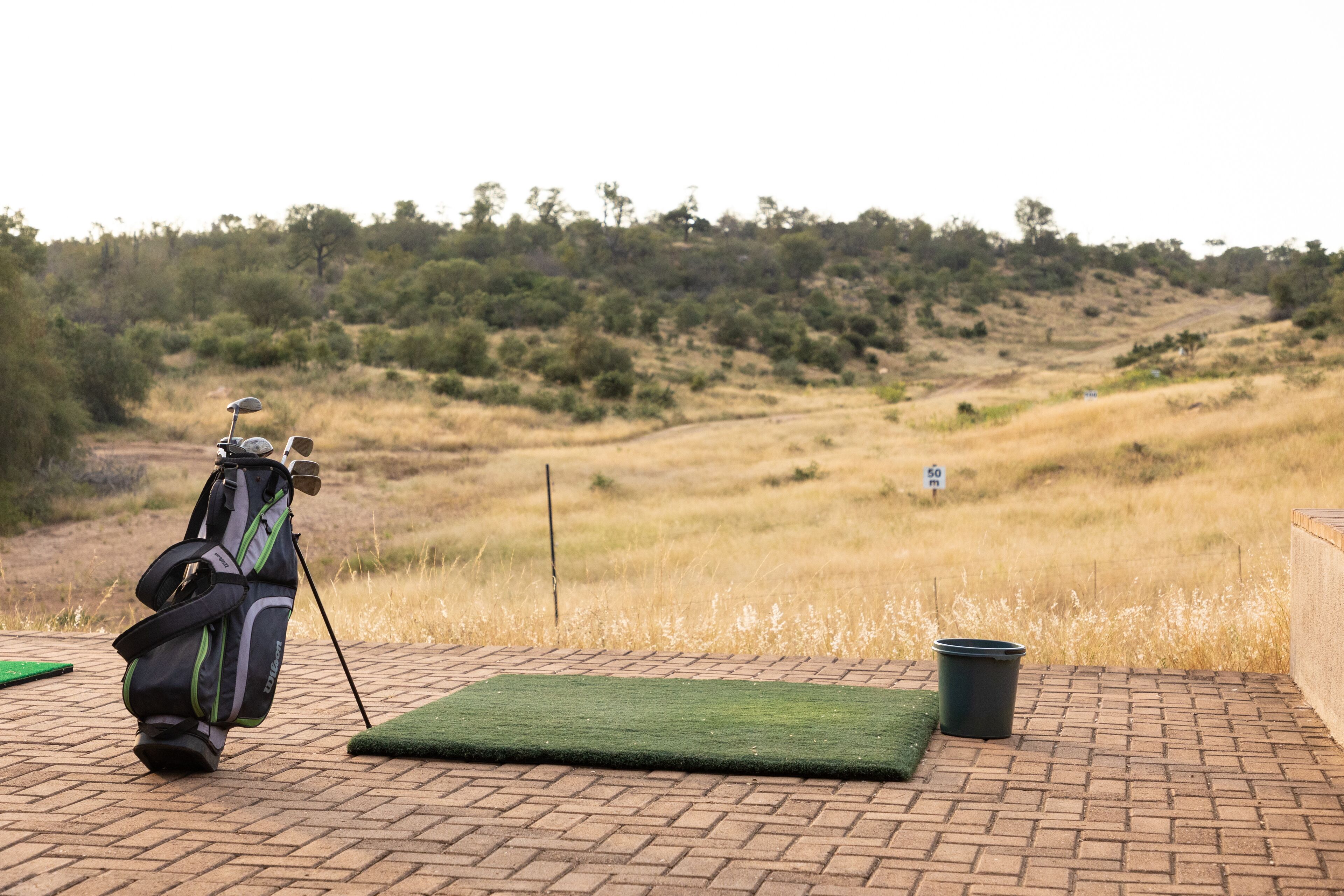 Un bolso de golf sobre una superficie de ladrillo con un tapete verde, un cubo negro y un cartel que indica una distancia de 50 metros.