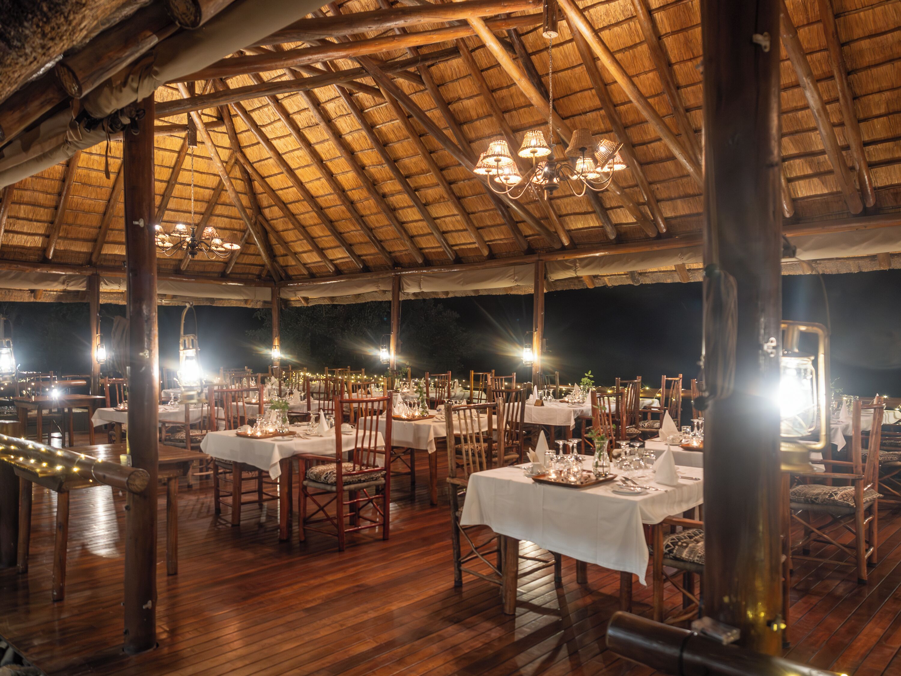 Un área de comedor al aire libre con mesas y sillas de madera, cubierta por un techo de paja e iluminada por luces colgantes.