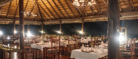 Un área de comedor al aire libre con mesas y sillas de madera, cubierta por un techo de paja e iluminada por luces colgantes.