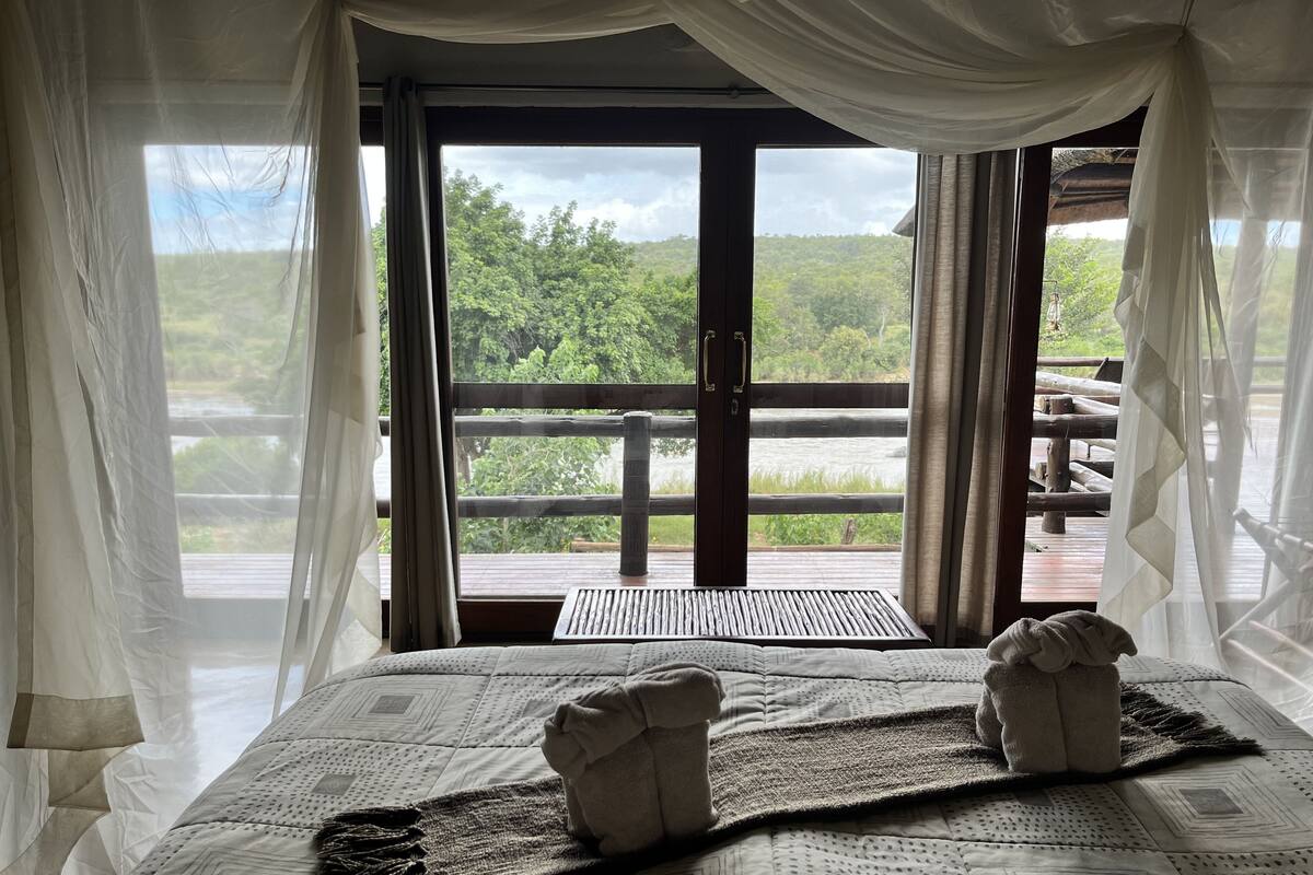 A bed with a view of a wooden deck and greenery, curtains, and a window with a view.