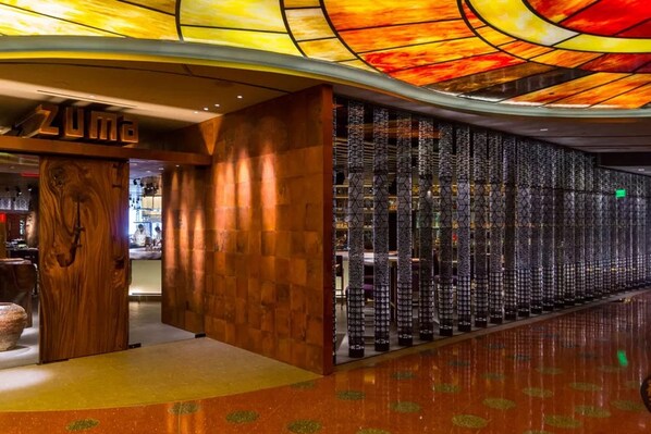 A hotel interior with a wooden reception desk, a wall of hanging luggage, and a ceiling with a stained glass design.