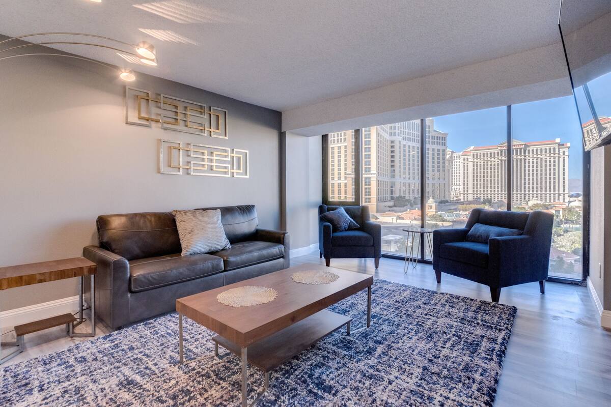 A modern living room with a leather sofa, two armchairs, a coffee table, and a large window offering a city view.