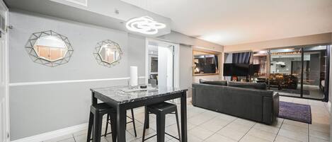 Una sala moderna con un sofá de color oscuro, una mesa de comedor con dos taburetes y una cocina visible a través de puertas de cristal.