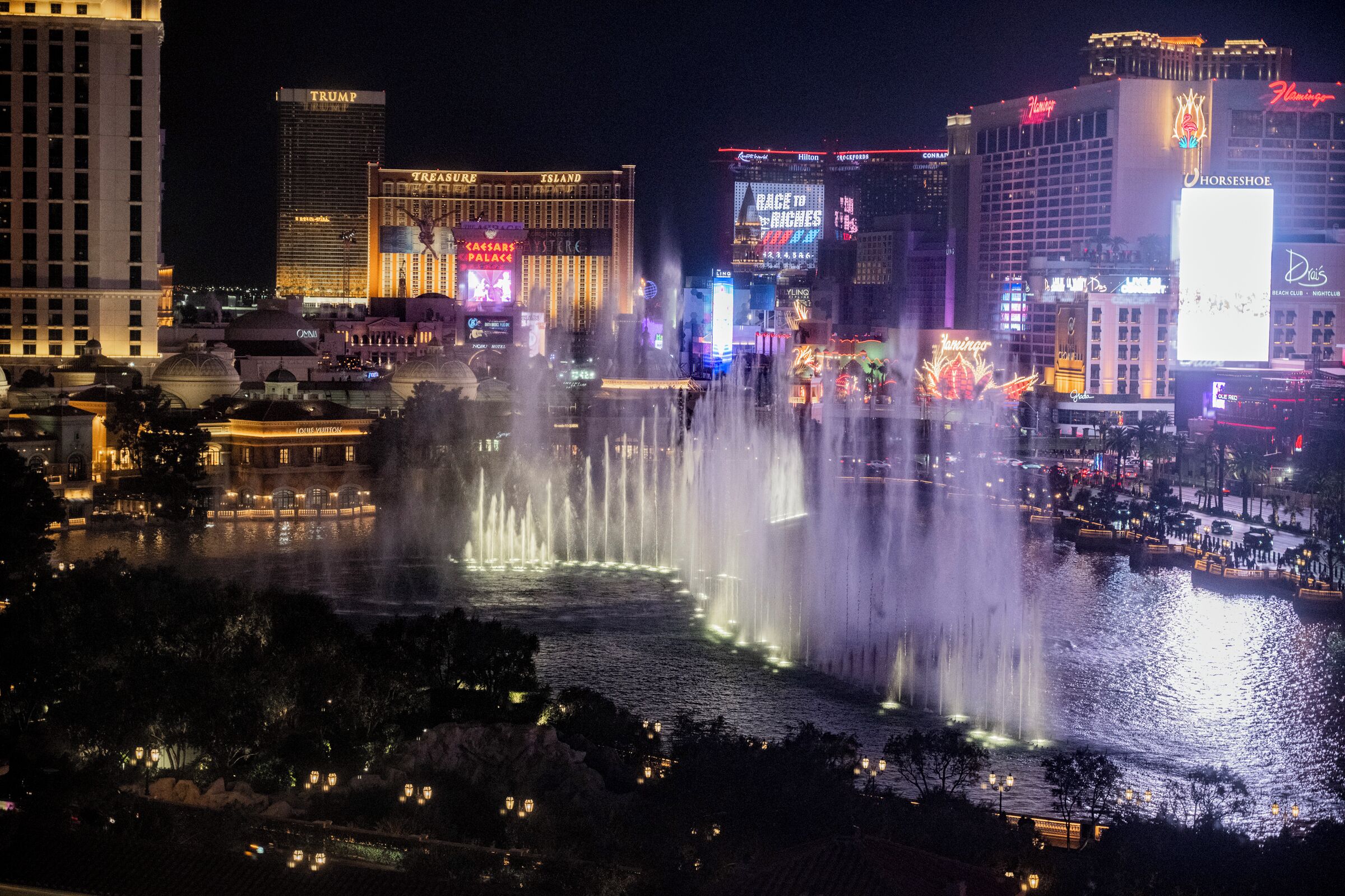 A nighttime cityscape with illuminated buildings and a water fountain display.