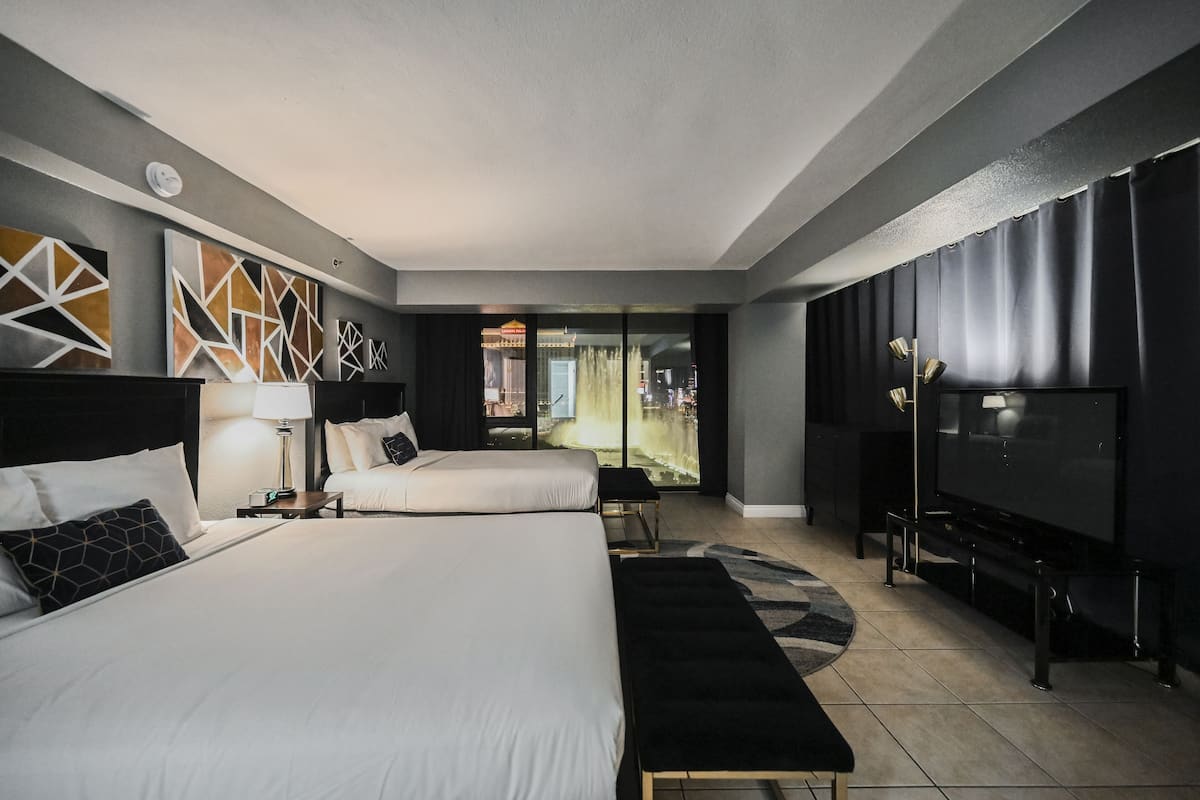 A hotel room with two beds, a TV, and a view of a cityscape through large windows.
