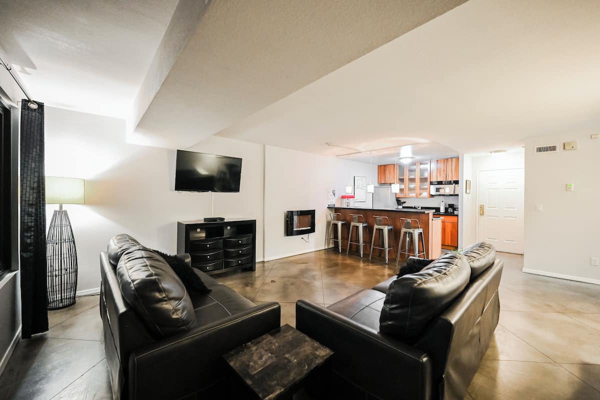 A spacious living area with a black leather sofa, a TV mounted on the wall, and a kitchen in the background.