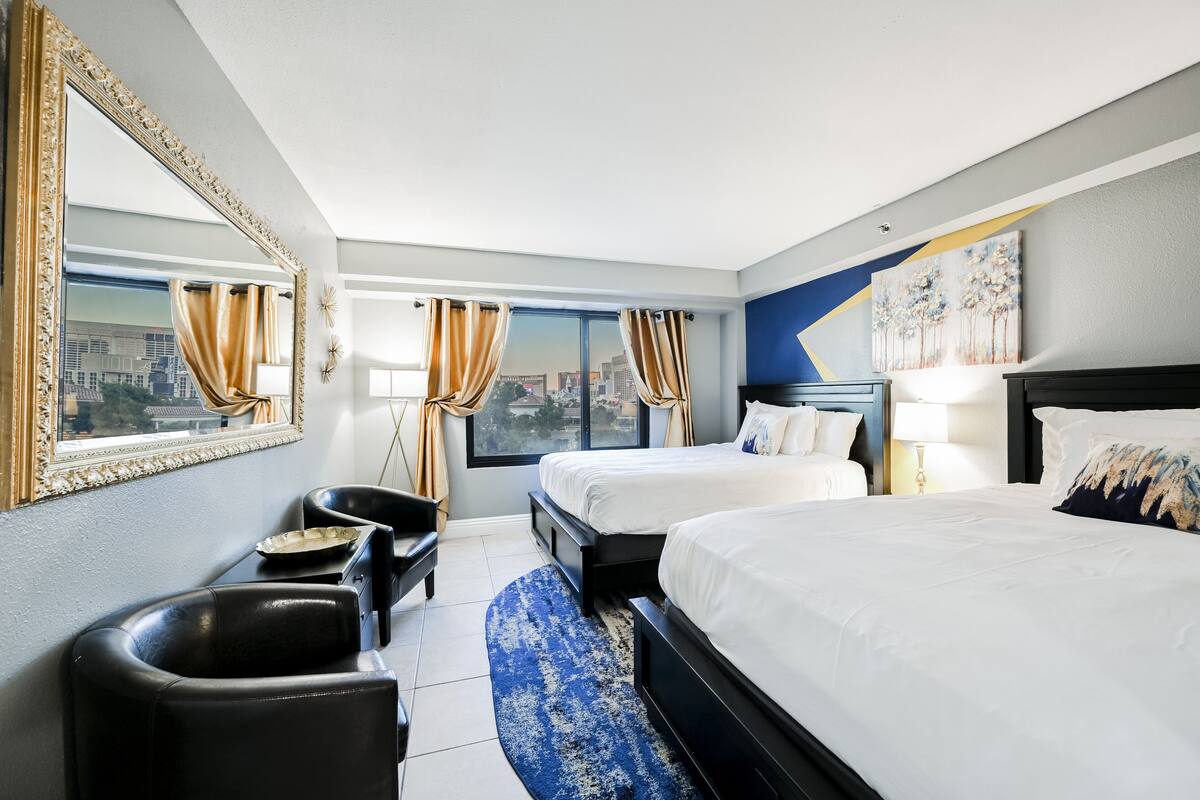 A hotel room with two beds, a large mirror, a view of the city, and a blue patterned rug.