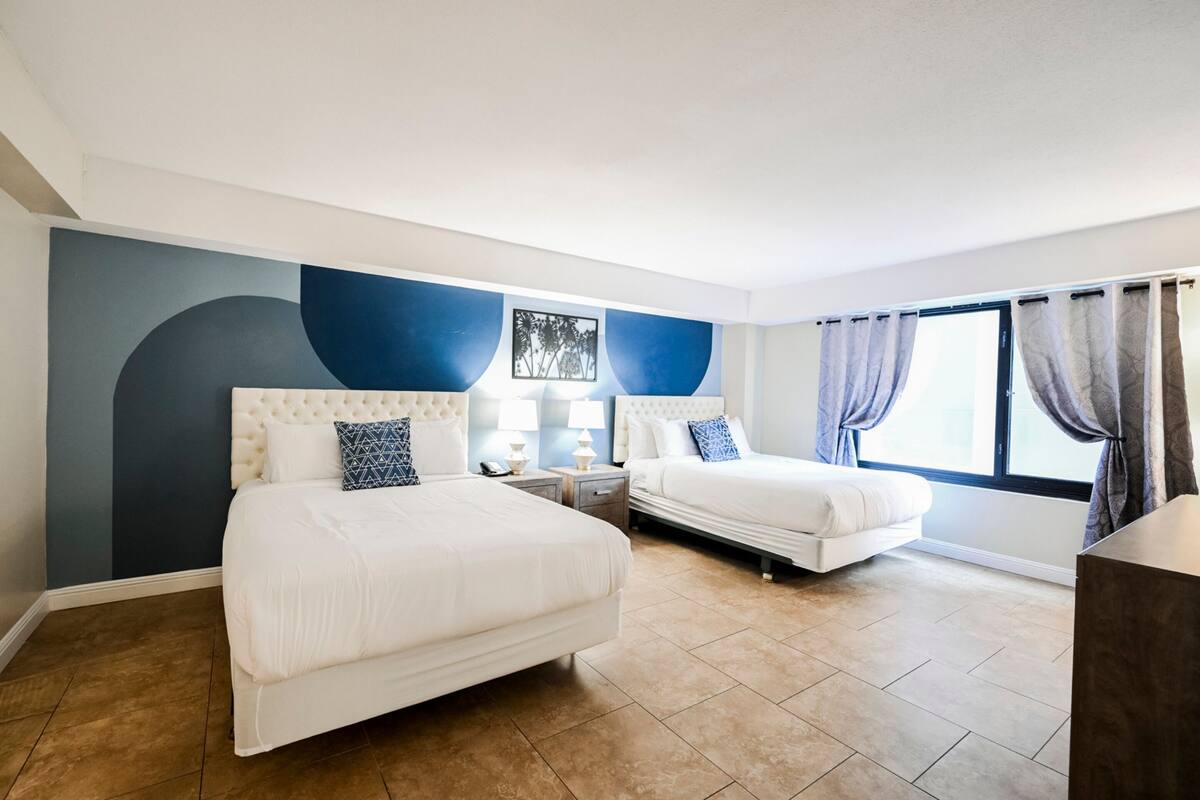 A hotel room with two beds, a large window with curtains, a wall-mounted artwork, and a nightstand with a lamp.