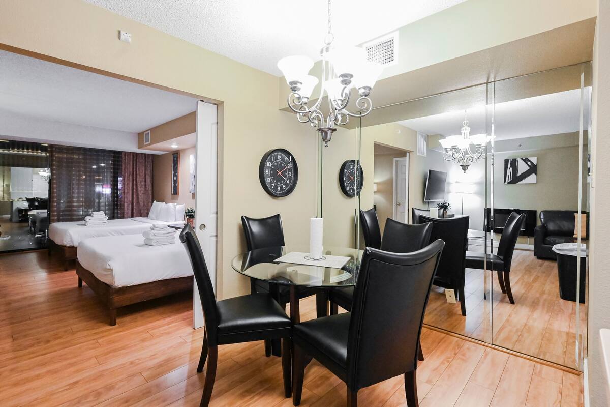 A hotel room with a bed, a glass dining table with chairs, a chandelier, and a large mirror reflecting the room.