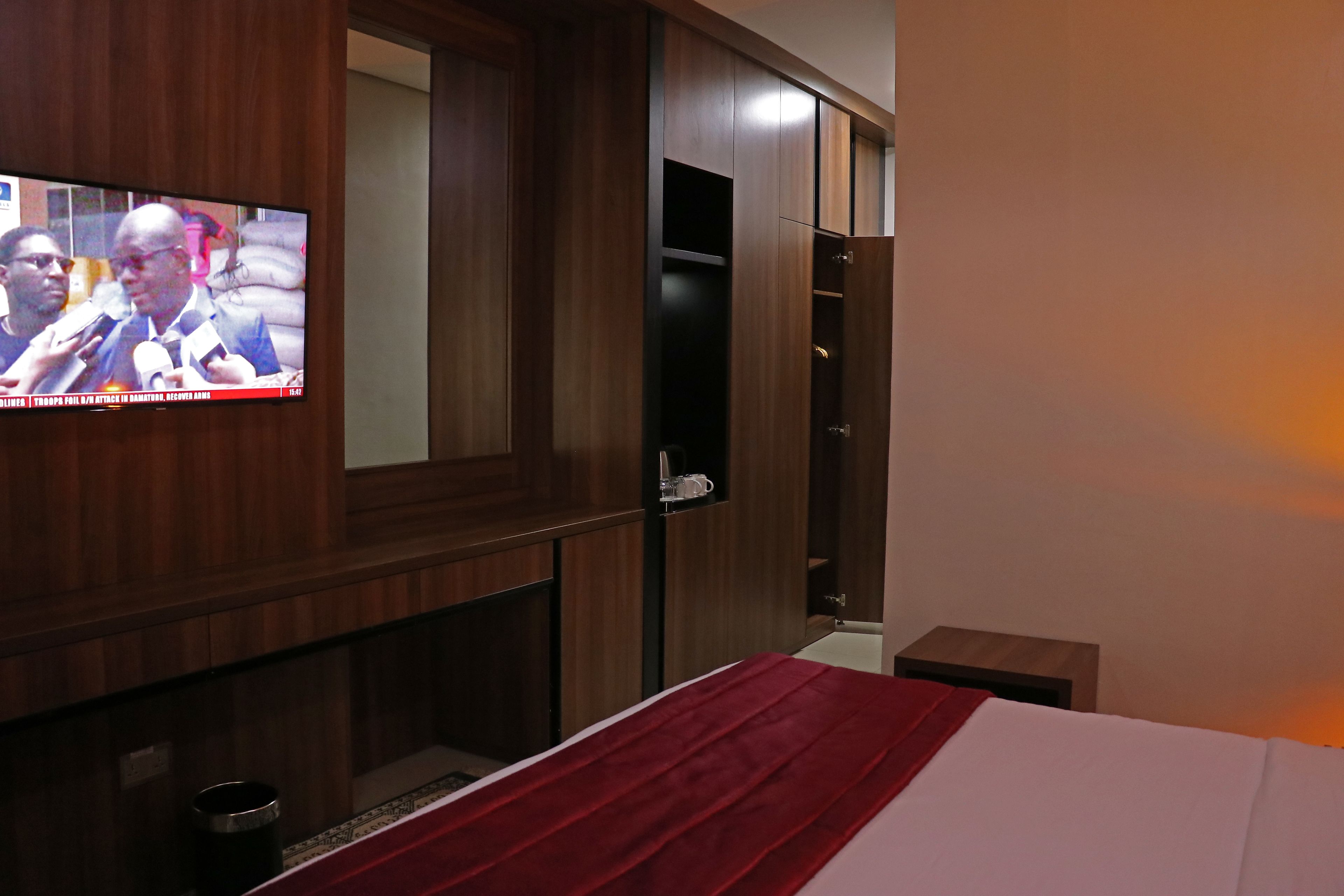 A hotel room with a television mounted on the wall, wooden wardrobe, and a bed with a red blanket.