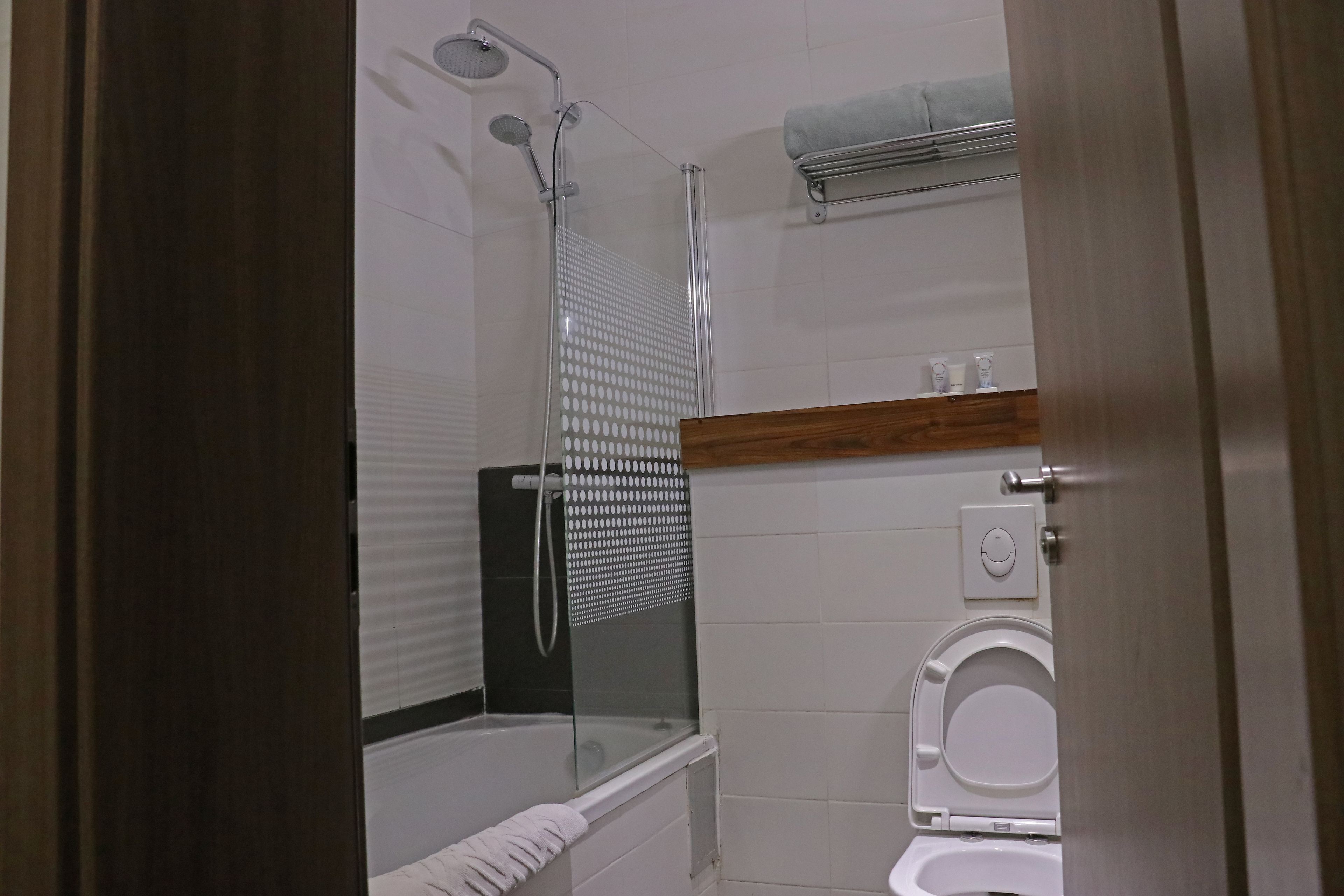 A bathroom with a glass-enclosed shower, a white toilet, and a wooden shelf above the toilet.