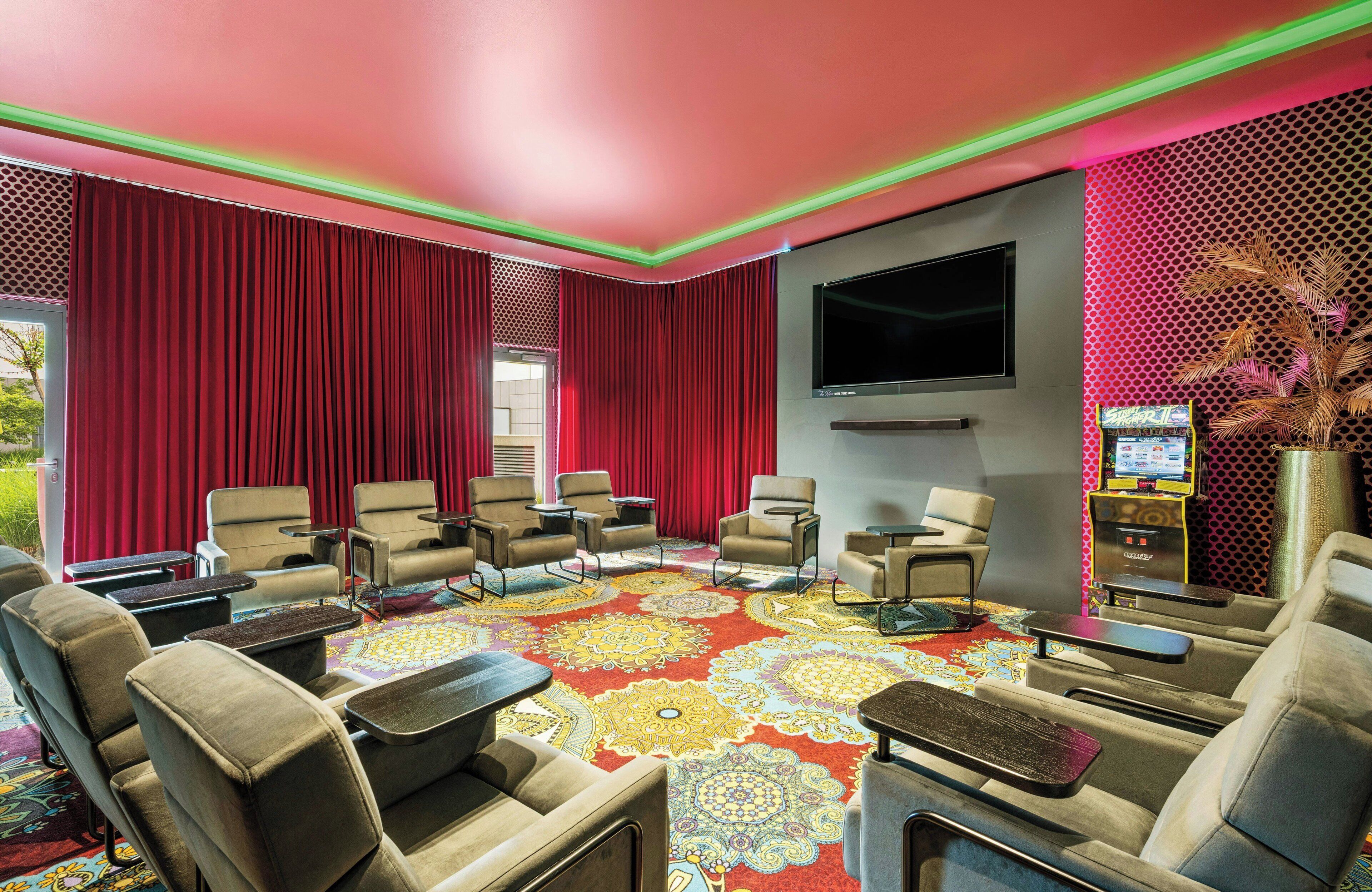 Una habitación con una cortina roja grande, un televisor de pantalla plana, varios sillones, una alfombra estampada y una pequeña mesa con una planta.