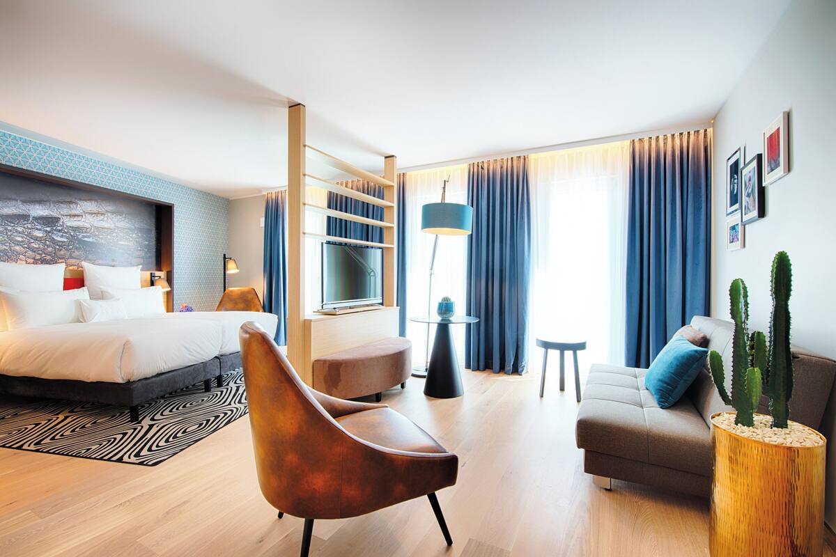 A modern hotel room with a bed, a sofa, a chair, a small table, a lamp, a cactus plant, and a large window with curtains.