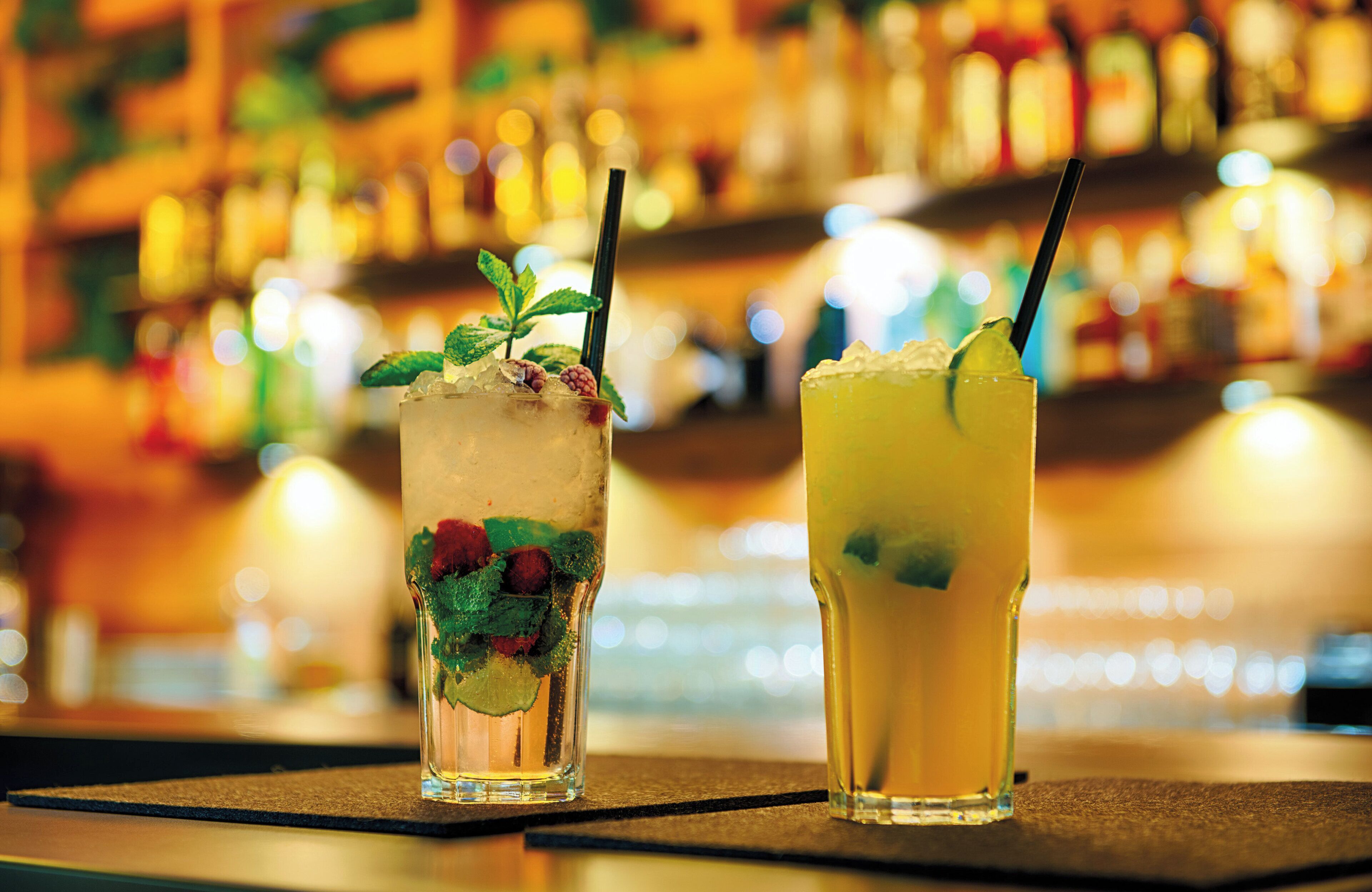 Two cocktails on a bar counter with a blurred background of bottles.