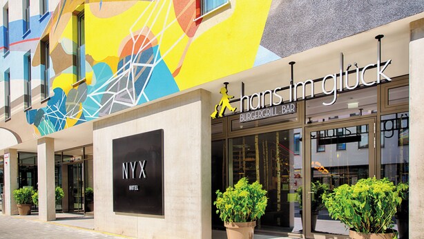 A hotel entrance with a colorful mural, a sign for 'Hans Im Glück Burger Grill Bar', and a 'NYX Hotel' sign. - NYX Hotel Munich by Leonardo Hotels (Munich)