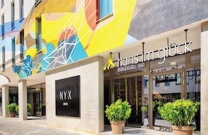 A hotel entrance with a colorful mural, a sign for 'Hans Im Glück Burger Grill Bar', and a 'NYX Hotel' sign.