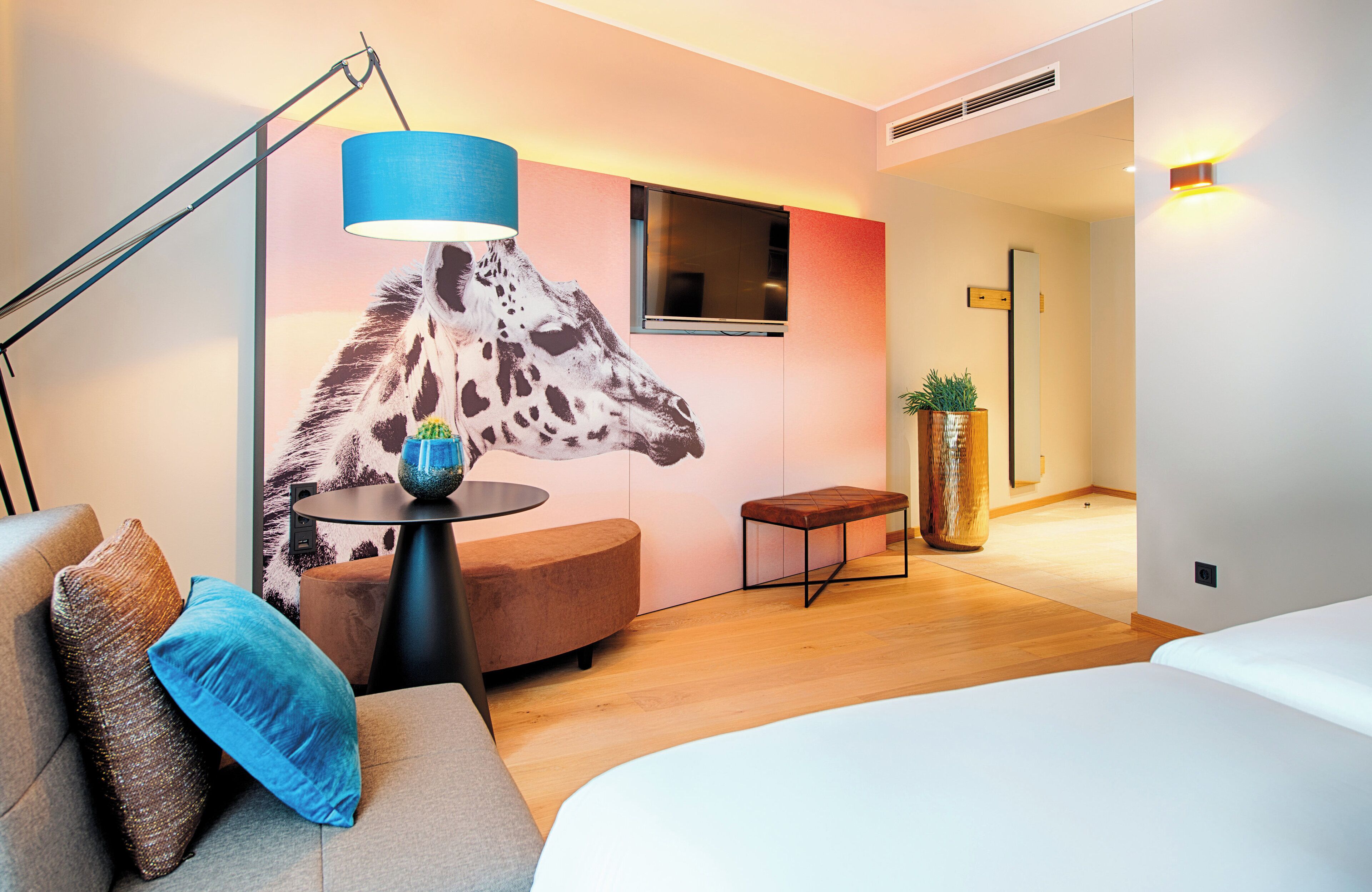A modern hotel room with a sofa, a bed, a desk, a giraffe mural, a TV, and a plant.