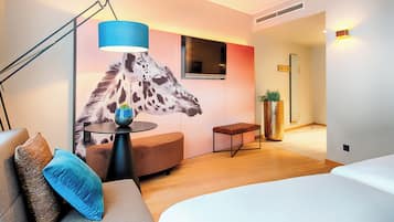 A modern hotel room with a sofa, a bed, a desk, a giraffe mural, a TV, and a plant.