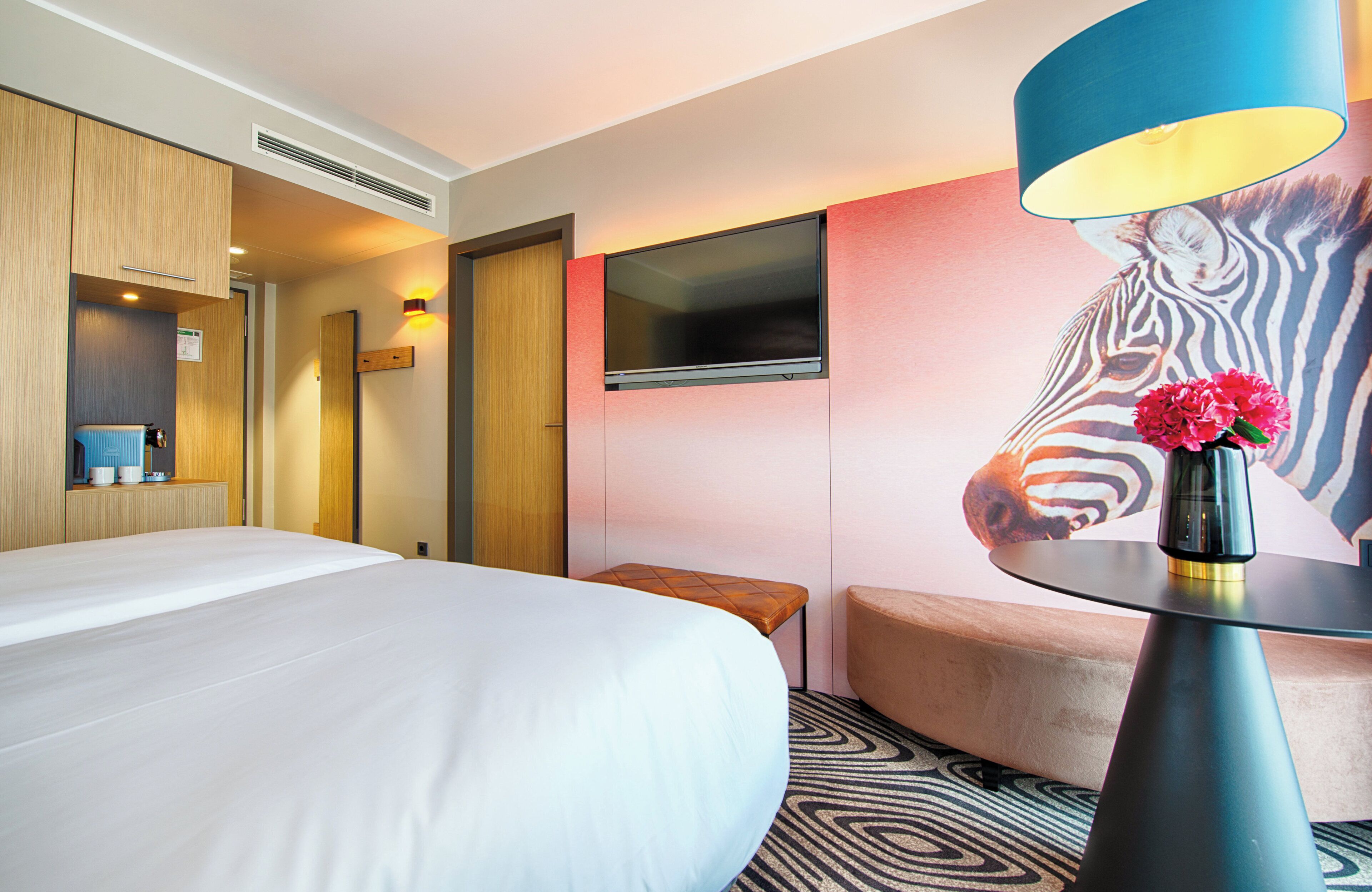 Une chambre d’hôtel avec un grand lit, un petit coin salon, un téléviseur fixé au mur et une lampe décorative agrémentée de fleurs.