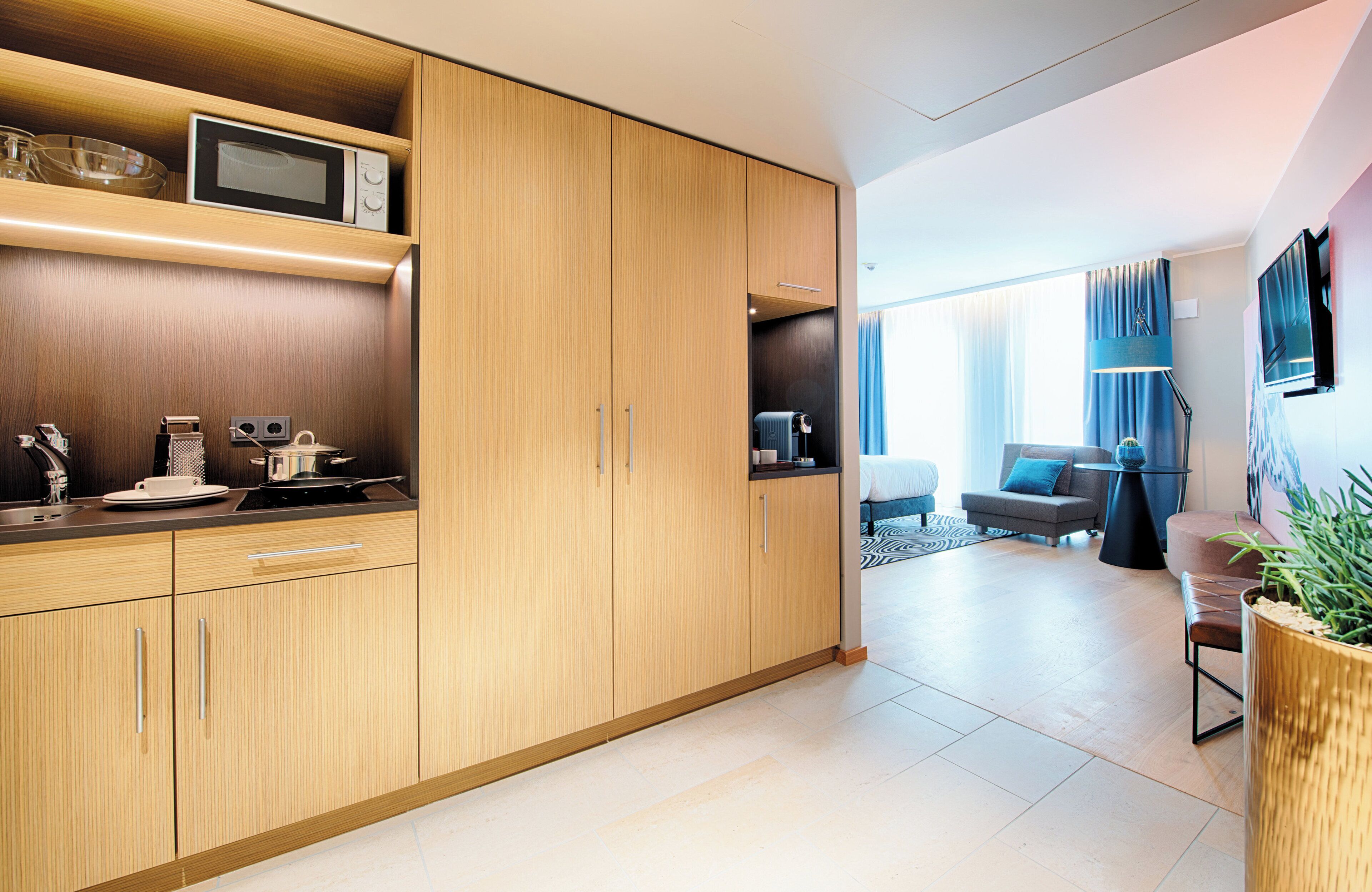 A modern kitchen with wooden cabinets, a microwave, and a coffee machine. There is a living area with a sofa and a TV.