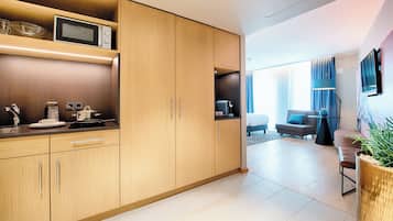 A modern kitchen with wooden cabinets, a microwave, and a coffee machine. There is a living area with a sofa and a TV.