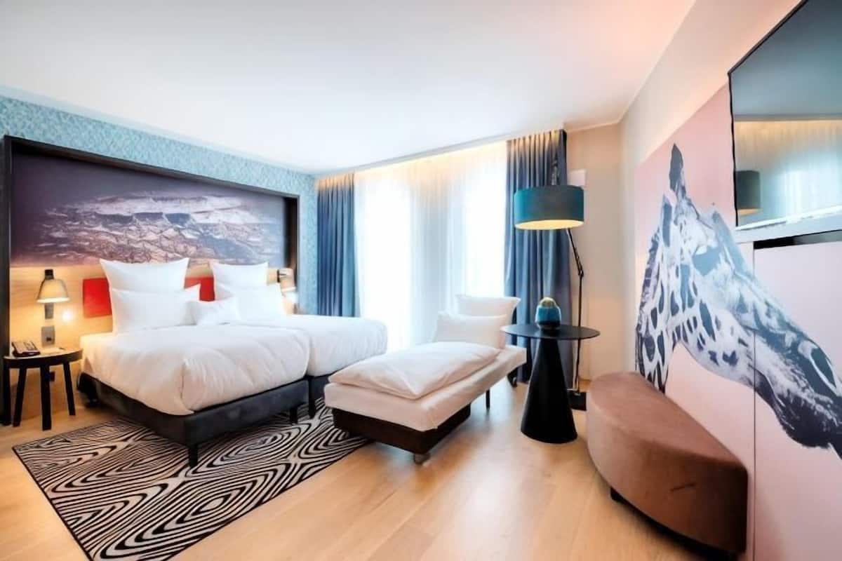 A modern hotel room with a double bed, a sofa, a desk, and a large window.